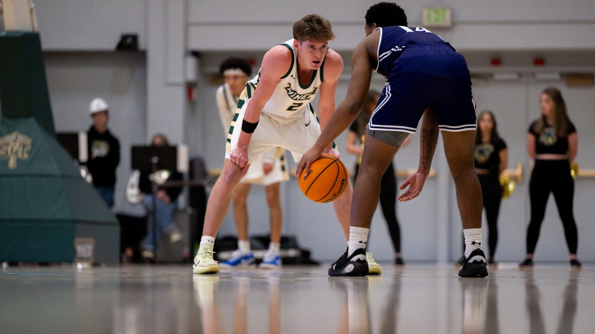 Missouri S&T men's basketball player Alex Benassi wearing a white jersey and standing in a defense stance with his hands on his knees while looking at a McKendree player who is dribbling the ball during a game in Gibson Arena 