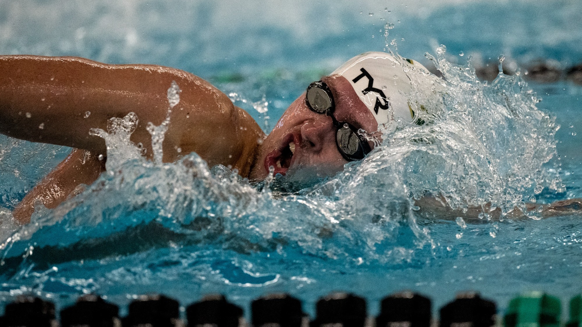 Missouri S&T swimmer Adam Wiedemeier is captured mid-race while swimming freestyle in an indoor pool. He wears a white Missouri S&T swim cap and dark swim goggles as he turns his head to the side to breathe, with his mouth open and face partially above the water’s surface. His left arm is extended forward beneath the water while his right arm recovers through the air, creating a splash that surrounds his head and shoulders. Lane lines are visible in the foreground, and the blue water fills the frame, emphasizing speed, motion, and competitive intensity during a live swim event.