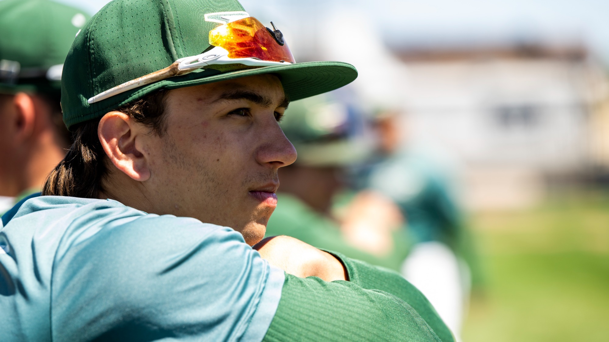 Missouri S&T baseball player Drew Baugus is shown in a close-up, side-profile view while leaning forward with his arms resting on the padded dugout railing. He wears a green Missouri S&T baseball cap with reflective sunglasses resting on the brim and a green long-sleeve undershirt beneath a short-sleeve team top. His expression is focused and contemplative as he looks out toward the field. The background is softly blurred, showing other teammates in green uniforms and a sunlit baseball field, emphasizing a live game or practice setting.