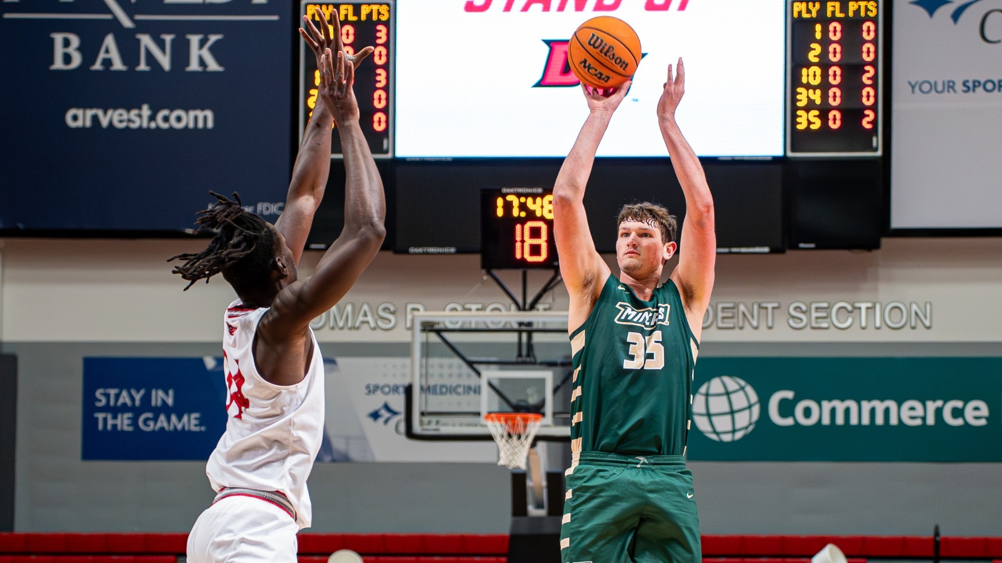 Zac Brown, a Missouri S&T men’s basketball player wearing a green jersey with the number 35, rises into a jump shot during a game inside an indoor arena. He holds the basketball above his head with both arms fully extended, focused on the basket. A defender in a white uniform jumps in front of him with arms raised in an attempt to block the shot. The basketball hoop and backboard are visible behind them, along with a digital scoreboard displaying the game clock and player statistics.