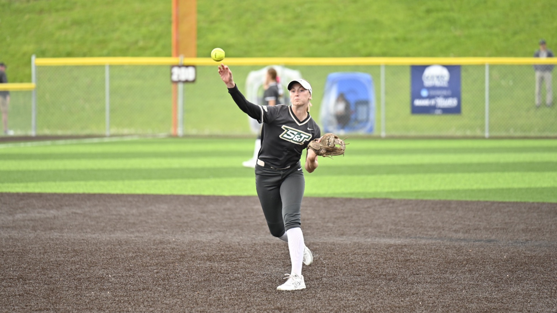 Trista Grobe, a Missouri S&T softball player, is photographed mid-throw on the infield during a game. She wears a black Missouri S&T jersey with white lettering, black pants, white knee-high socks, and a white visor. Her right arm is extended forward as she releases a yellow softball, while her left hand holds a glove close to her body. She is positioned on the dark turf infield with green outfield grass visible behind her, along with a chain-link fence, an outfield distance marker, and blurred figures in the background.