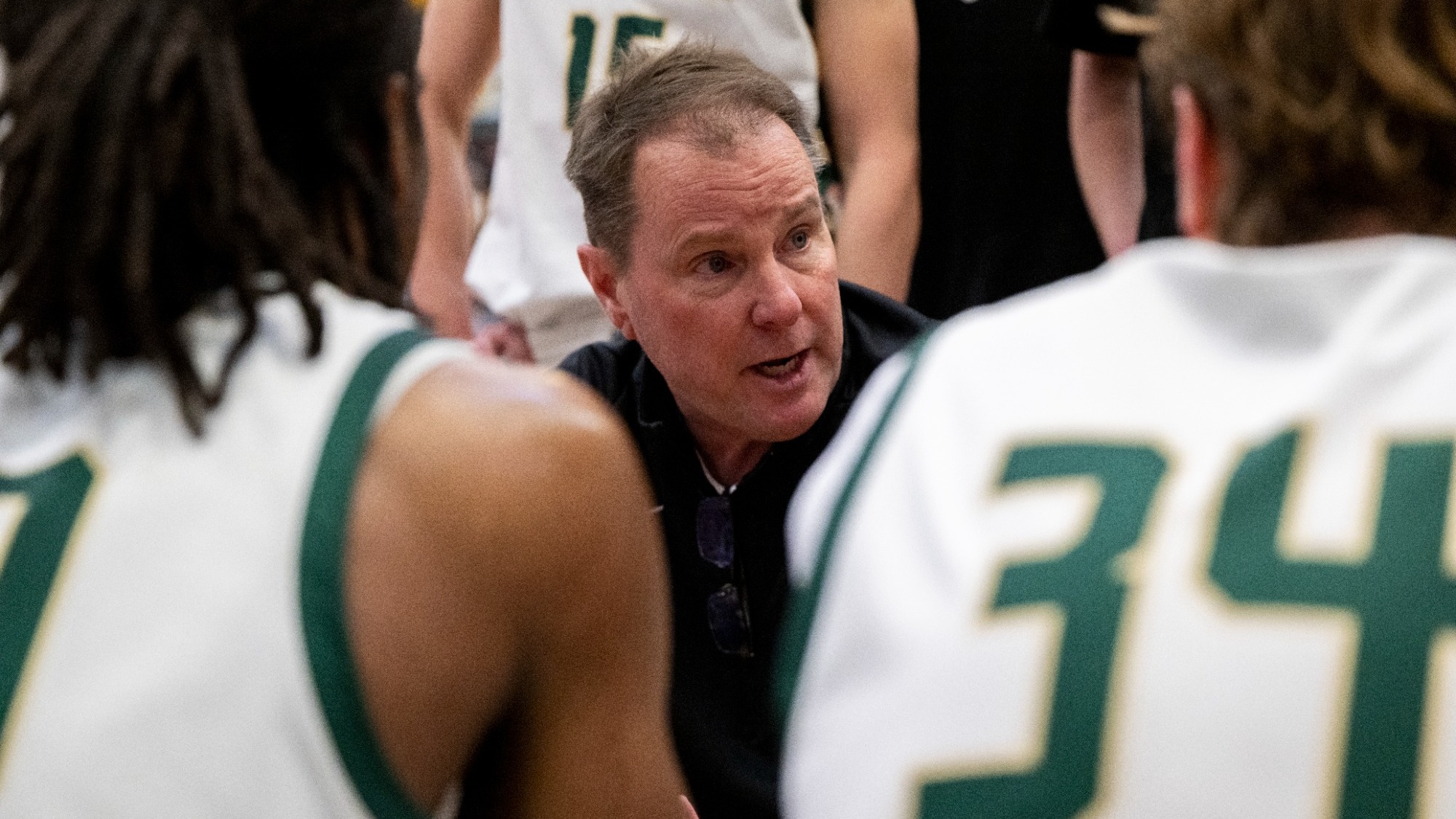 Photo of Head Coach Bill Walker wearing a black jacket and addressing his team during a timeout with Missouri S&T men's basketball players wearing white jerseys visible in the foreground and background