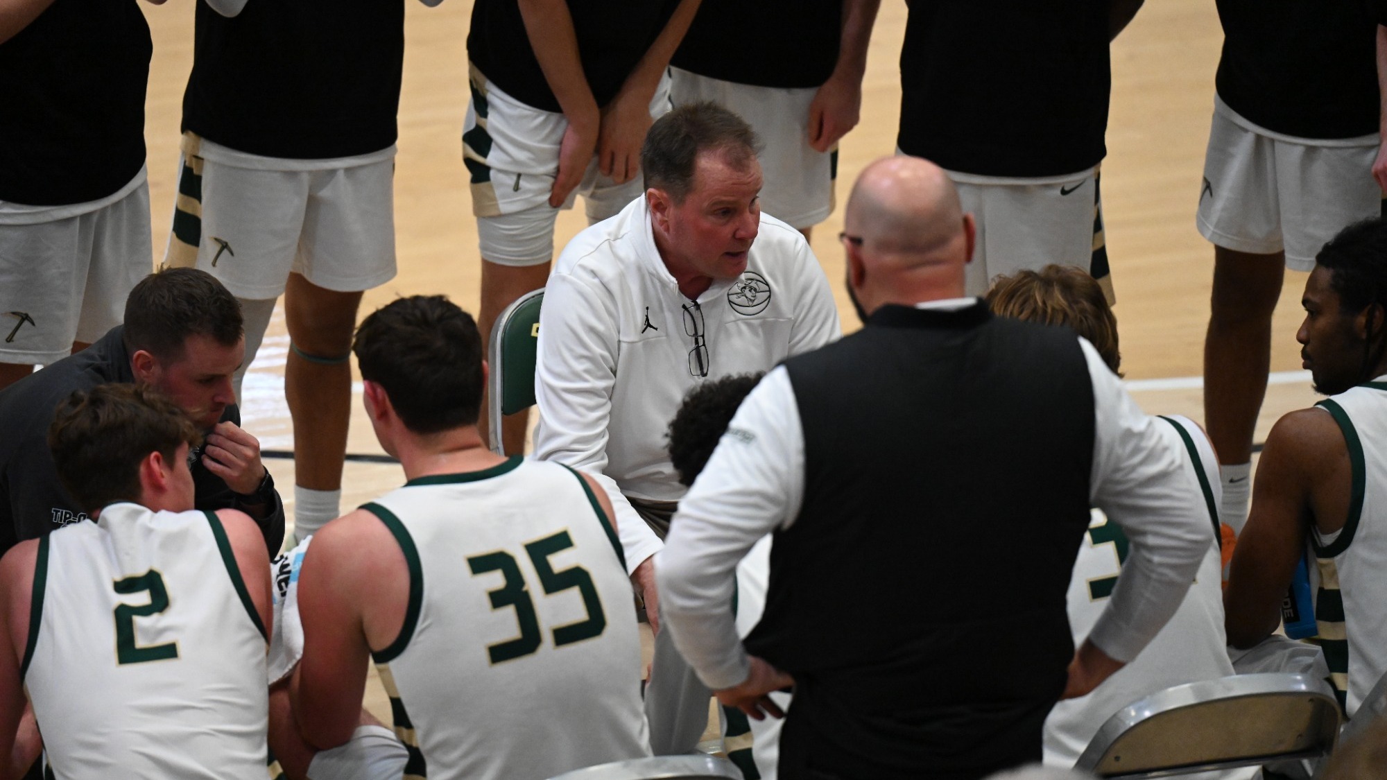 A photo of the Missouri  S&T Men's Basketball team sitting on the bench while Coach Bill Walker sits in front of them and talks to them