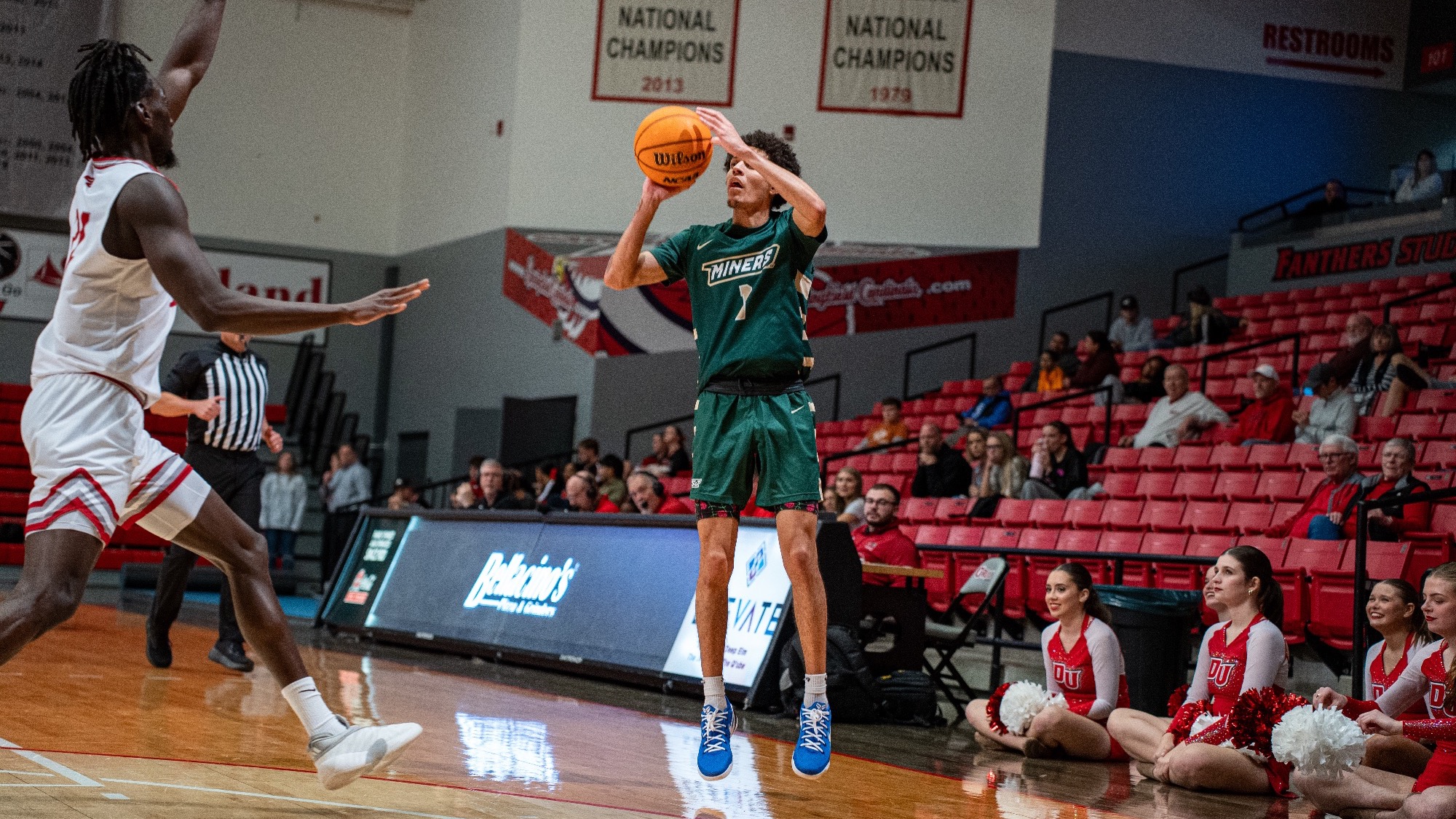 Missouri S&T men’s basketball player Blaise Beauchamp wearing a green jersey and jumping to shoot a three point shot while a Drury defender runs toward him to try and prevent the shot during a game at Drury University