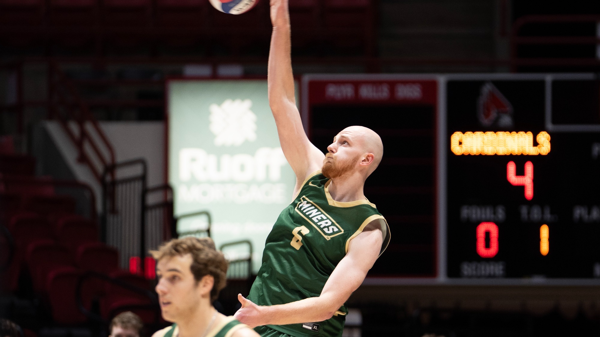 Missouri S&T Miners volleyball player Caleb Rohlwing jumps for a serve. He is reaching high with his right arm to serve the ball against a blurred background of a scoreboard and arena seating.
