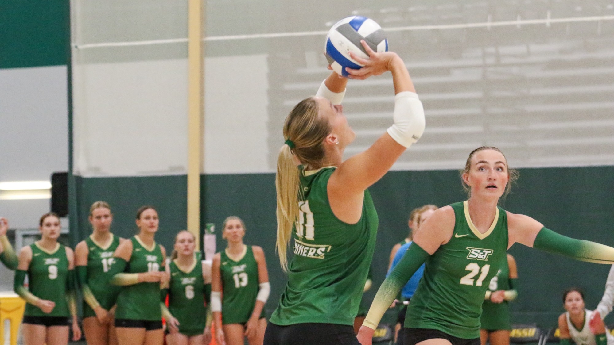 Maggie Miller of Missouri S&T sets a volleyball with both hands above her head during a match, with a teammate positioned beside her and other Miners players watching from the court and sideline.