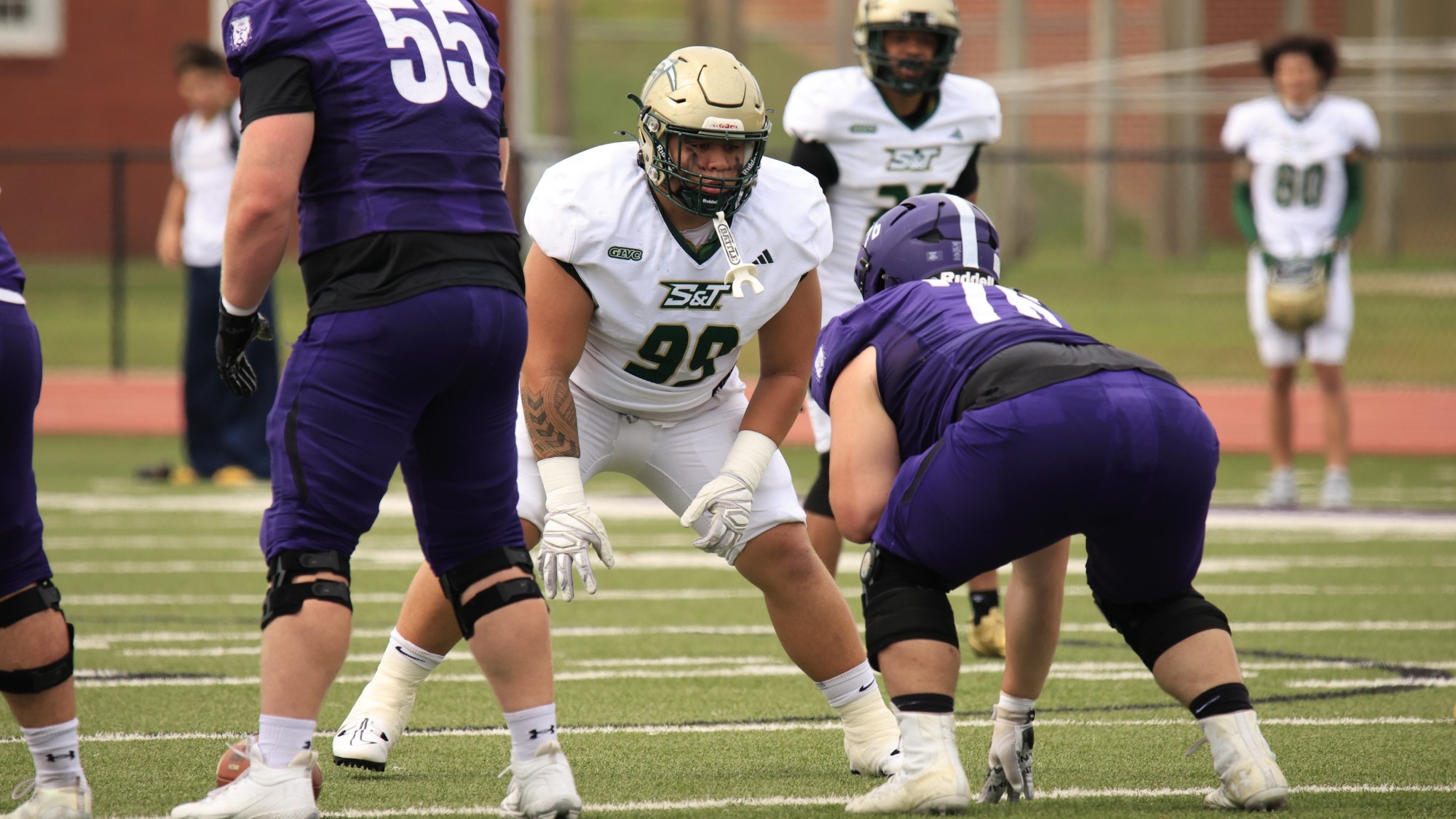 Mana Faagutu (#99) of Missouri S&T lines up as a defensive lineman against an opposing offensive line during a football game. He is crouched in a defensive stance between two opposing players in purple uniforms.