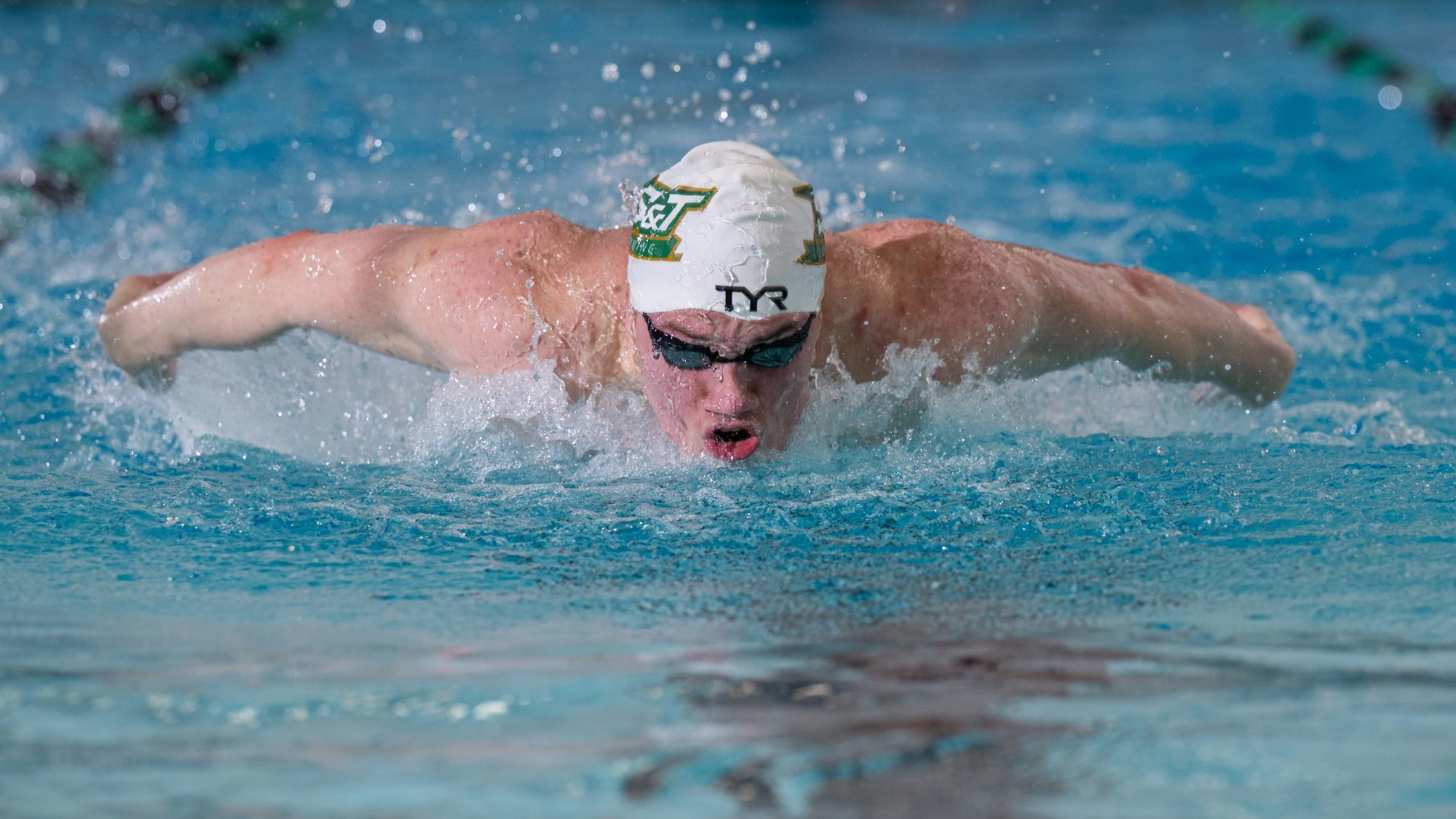 Oscar Iden of Missouri S&T swimming photographed doing the butterfly stroke in the S&T pool while wearing goggles and white swim cap with the Missouri S&T logo on it
