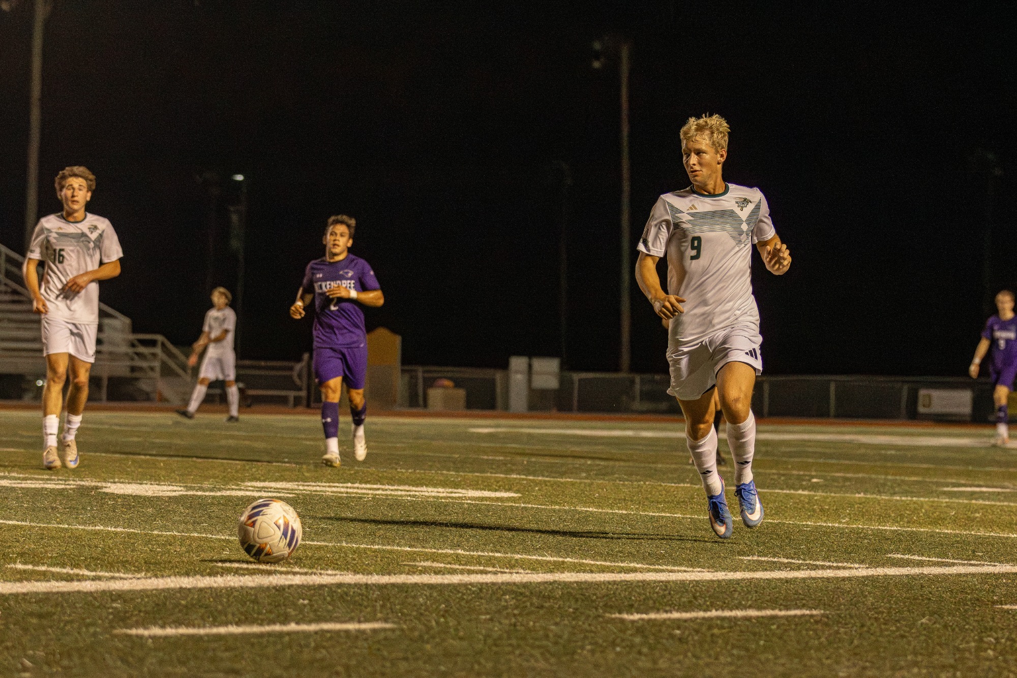 Mike Banasik in a white uniform jogging after the soccer ball on a grass field at night. 
