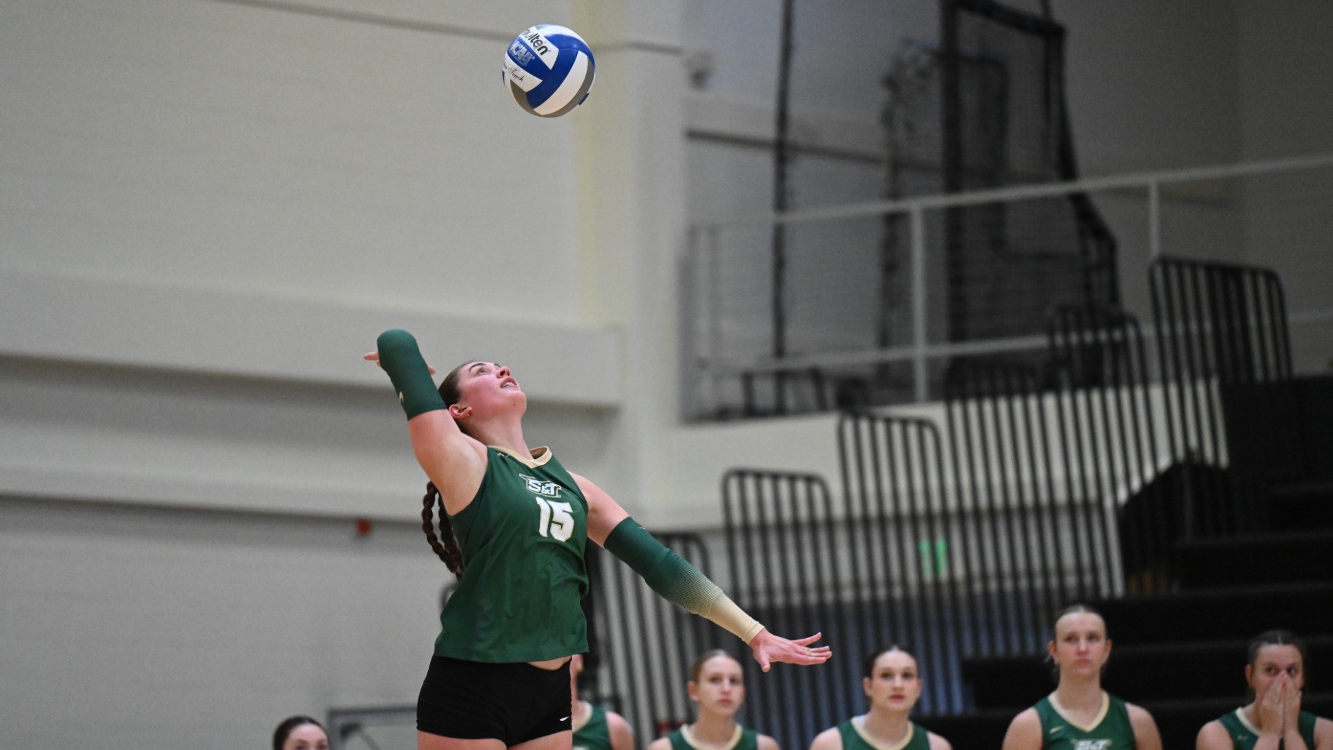 Elly Decker of Missouri S&T women's volleyball photographed midserve, jumping in the air and swinging her arm at a volleyball while wearing a green jersey
