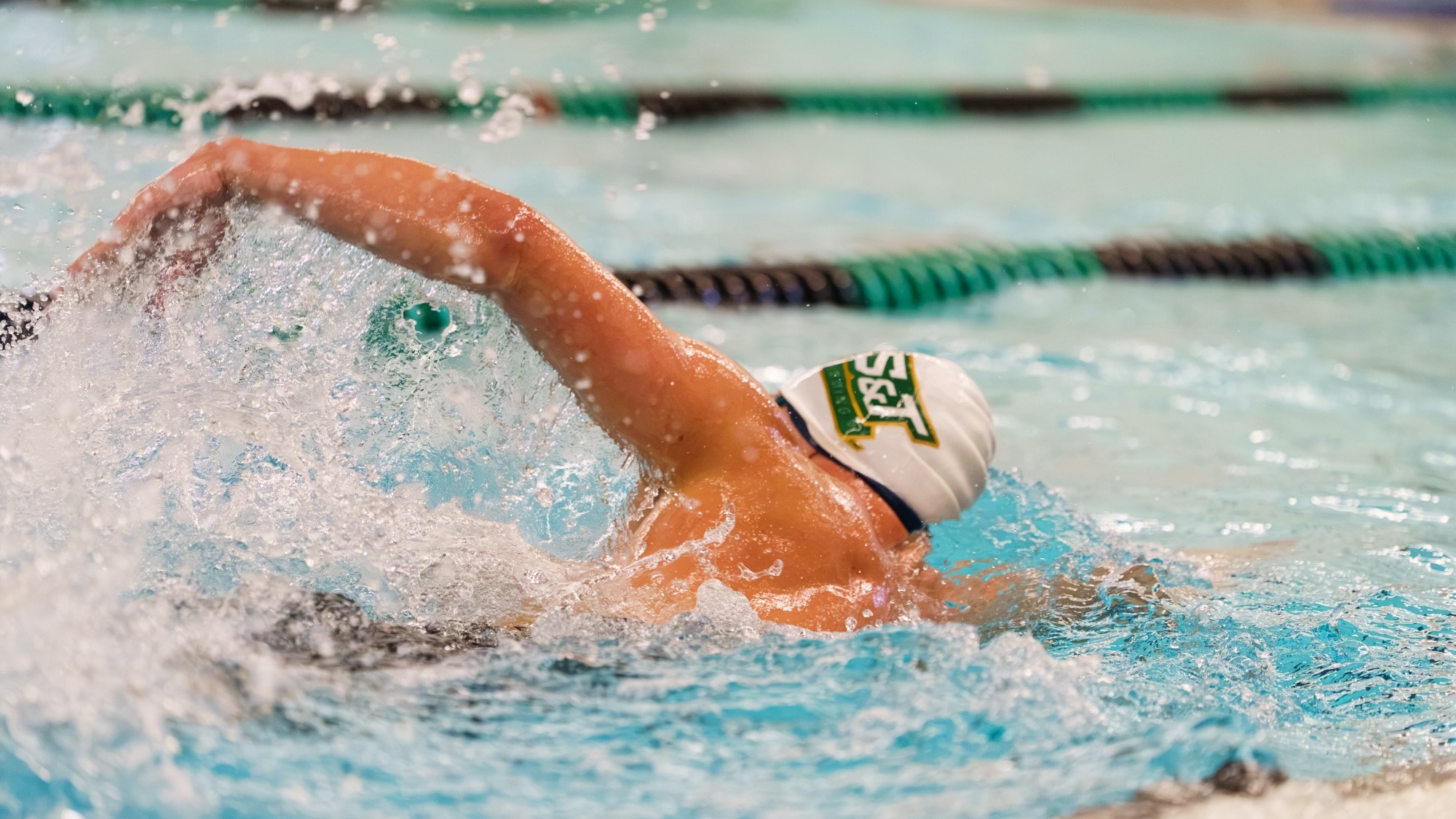 Missouri S&T Swimmer photographed during a freestyle event and wearing a white swim cap with the Missouri S&T logo on it