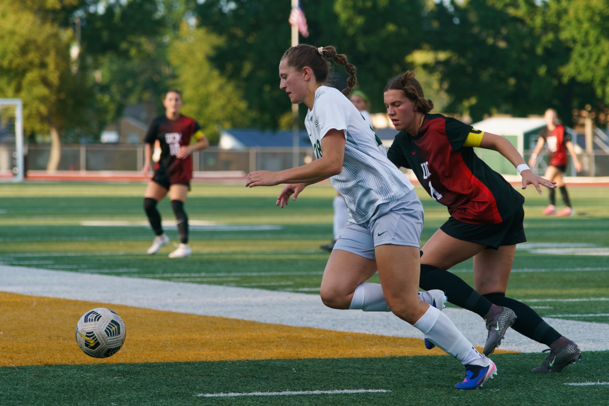 Andriana Benassi leaning forward dribbling a ball away from a defender. She is in a white jersey and white shorts