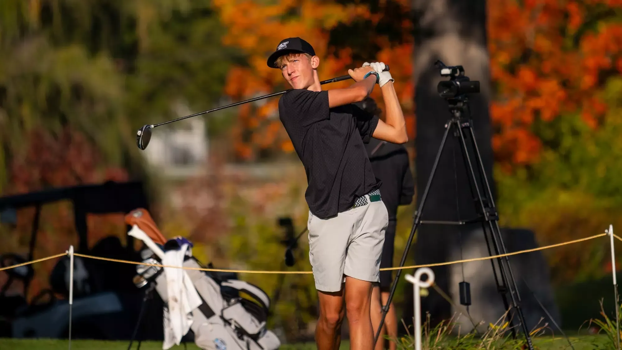 Lindahl of the Missouri S&T Men's Golf Team photographed wearing a black polo and white golfers shorts with a black baseball cap. He is mid-follow through on his swing during a tee shot at Findlay