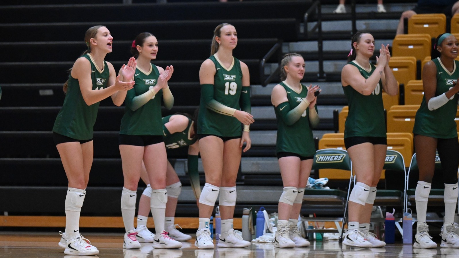 Missouri S&T Women's Volleyball team photographed cheering from the bench while wearing green uniforms