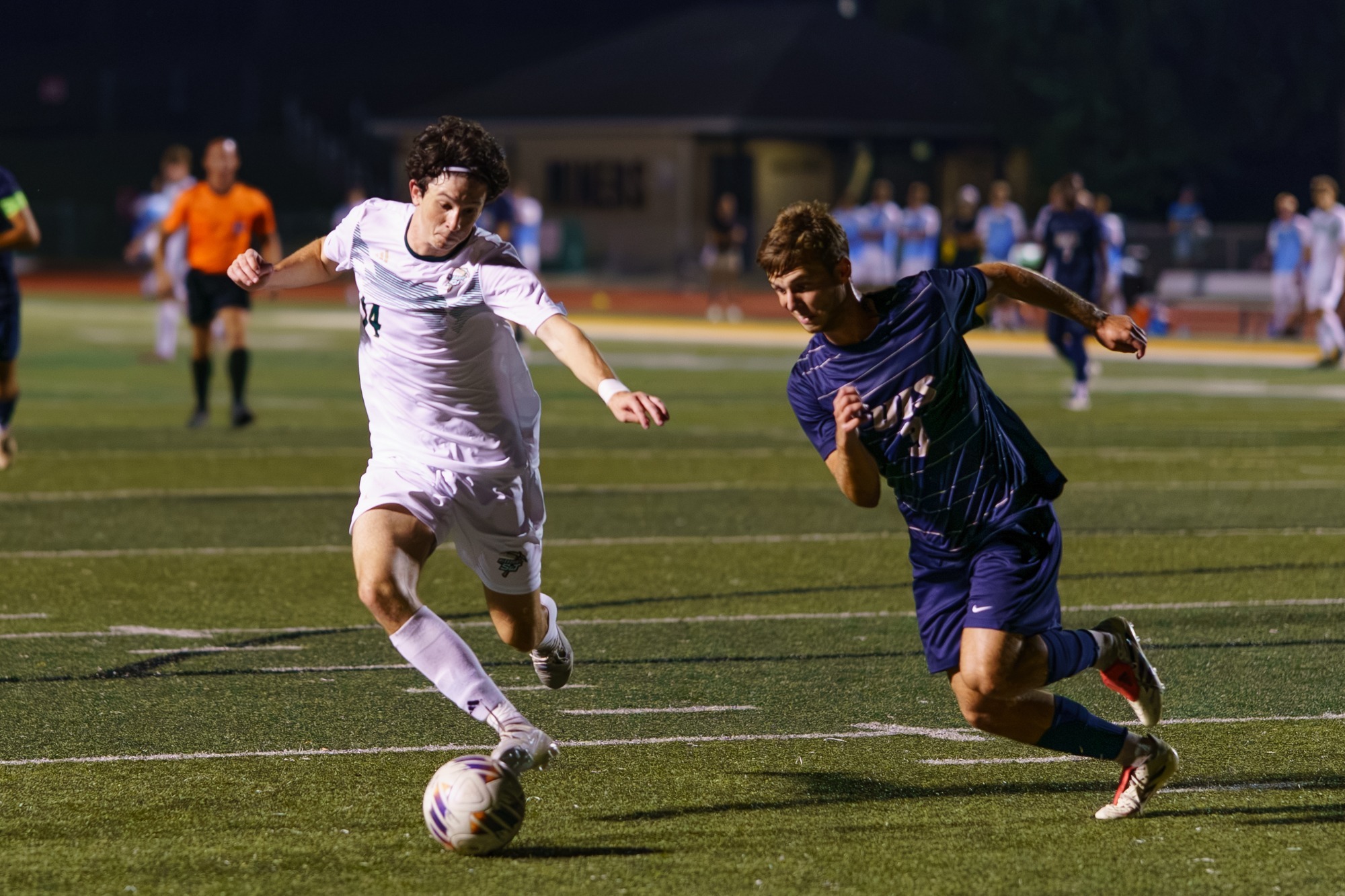 Caleb Lepant in a white jersey and white shorts dribbling the ball away and dodging a defender who is wearing a purple jersey and purple shorts. 