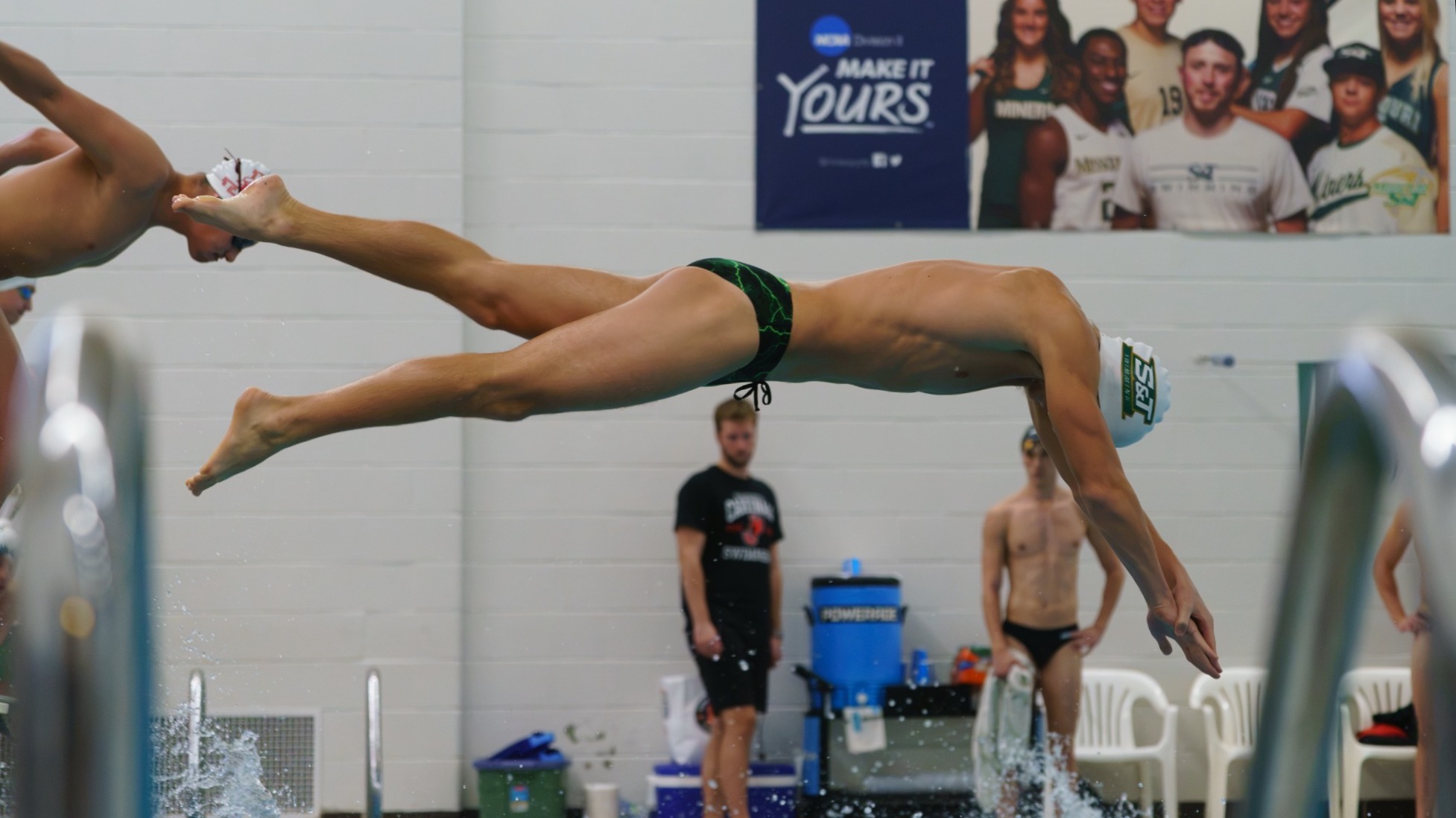Missouri S&T Swimmer photographed Diving into the pool during a meet. He is wearing a white swim cap and green speedo