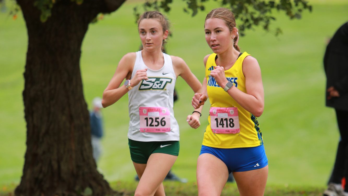Aurora Amason running in a white jersey and running tag alongside a runner in a yellow jersey and blue shorts