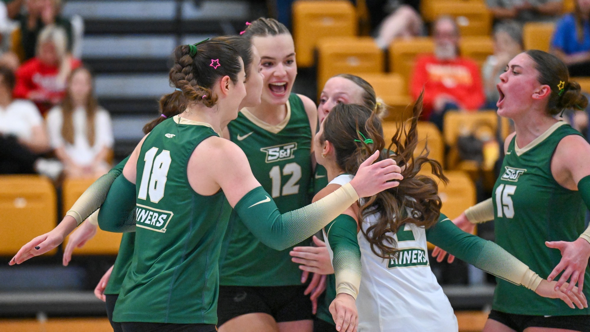 Women's Volleyball Team Photographed huddling and cheering during a match in Gibson. They are all wearing green and white jerseys.