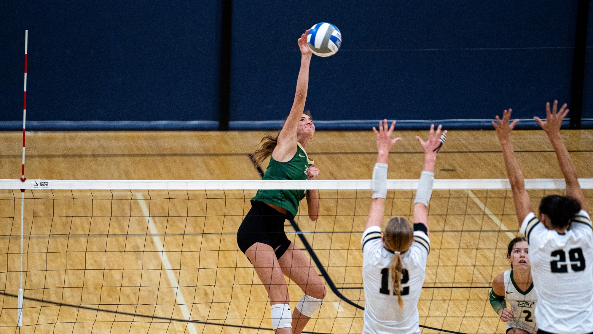 Addie Schoenberger of Missouri S&T women's volleyball photographed jumping above the net and attacking a volleyball while wearing a green uniform.