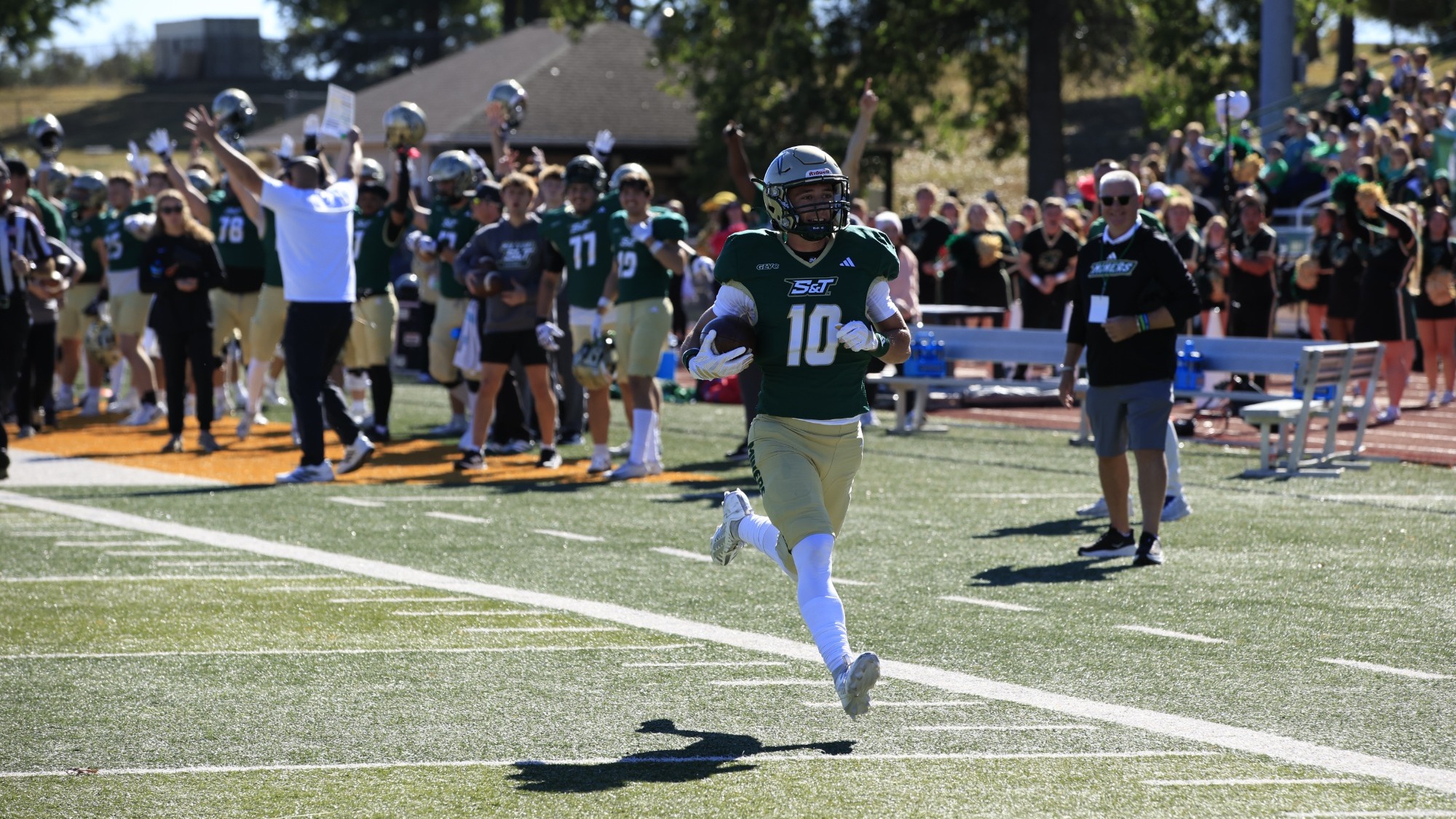 Tyler Gioia of Missouri S&T football photographed wearing a green and gold uniform with a gold helmet. He is holding the football and running into the endzone for a touchdown while fans and teammates cheer in the background