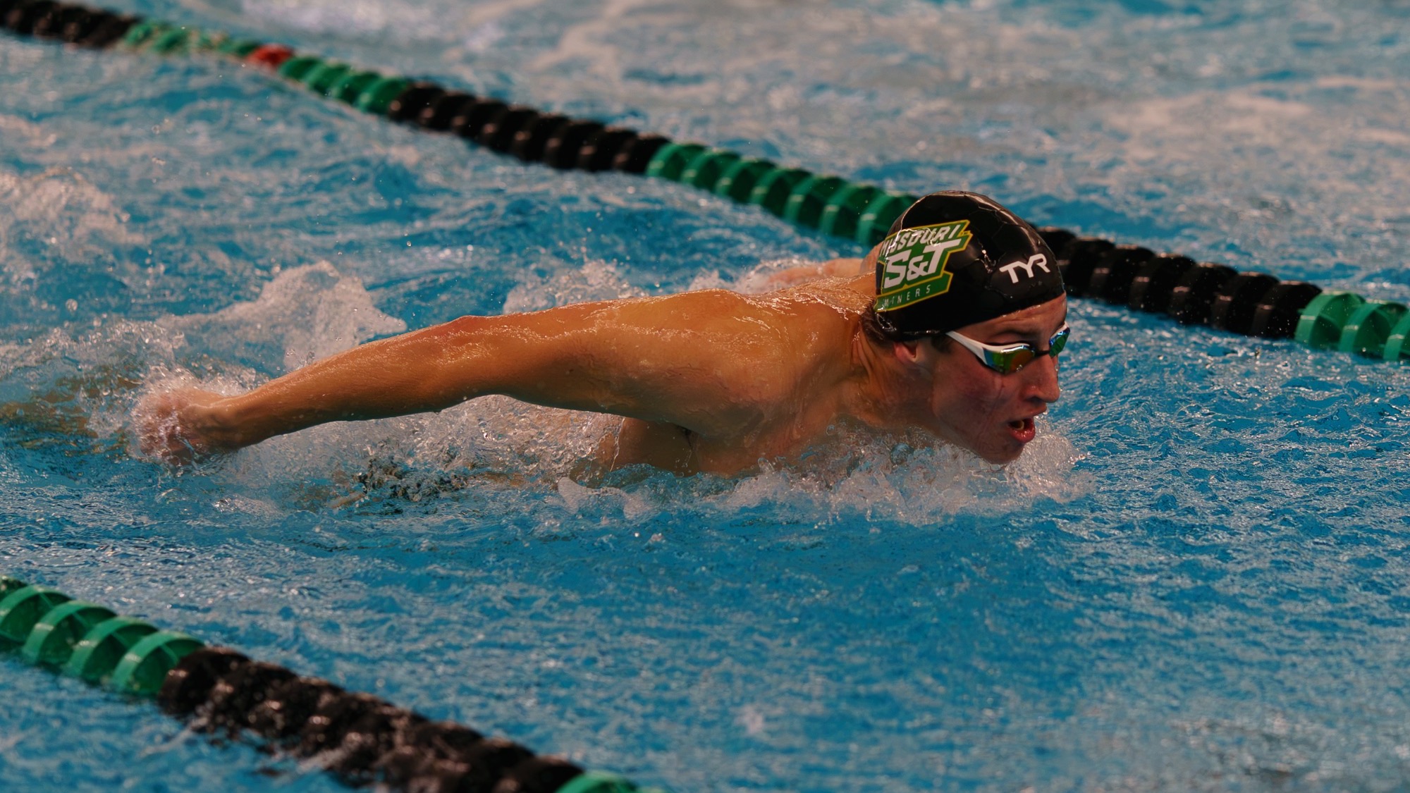 Otto Lovdahl of Missouri S&T swimming photographed doing the butterfly stroke during a meet in the S&T pool. He wears a black swim cap with the Missouri S&T logo on it