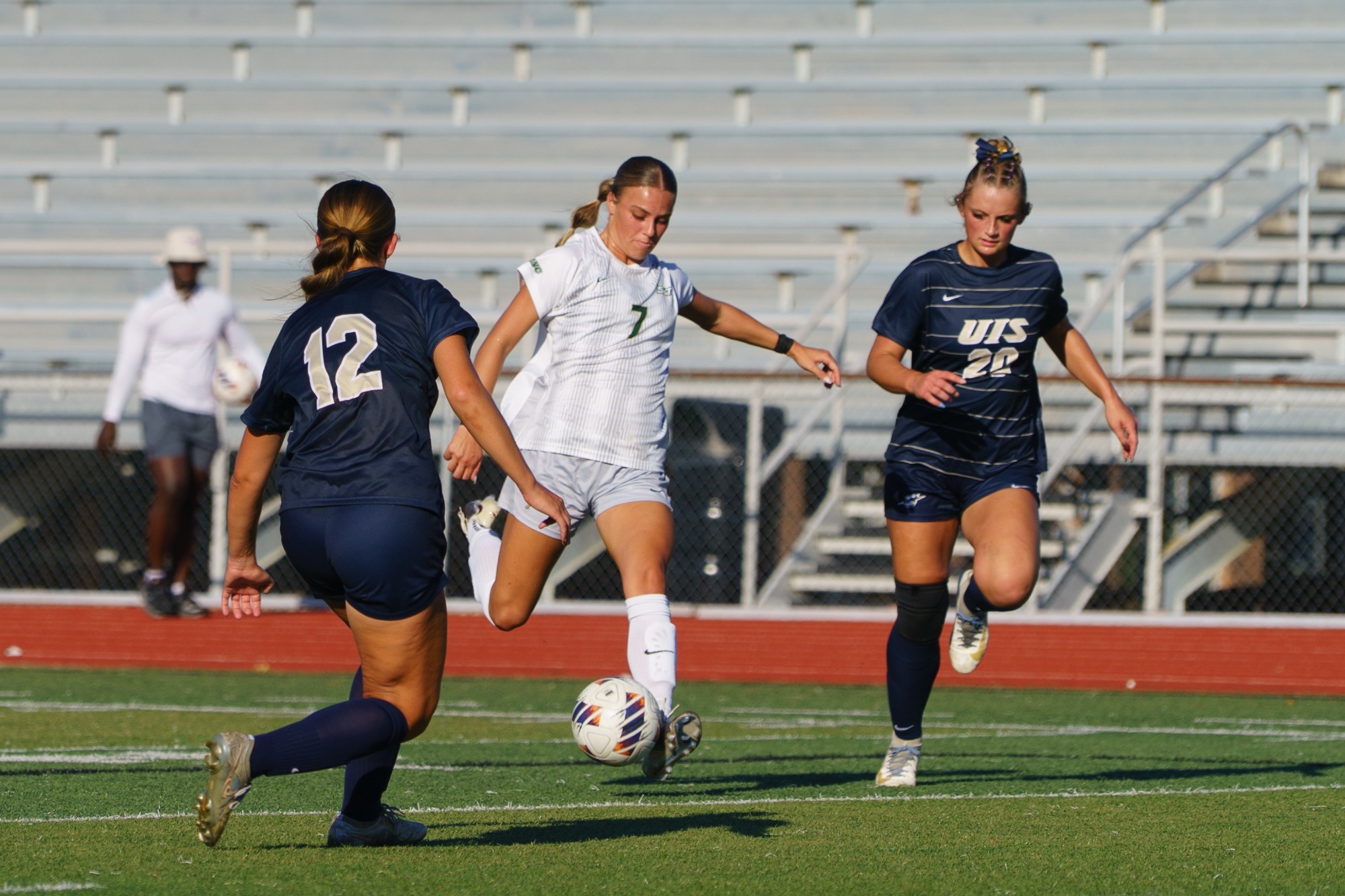 Haylee Prime for Missouri S&T women's soccer winding up to kick the soccer ball. She is wearing a white jersey and white shorts with UIS defenders in navy blue jerseys and navy blue shorts on each side of her.