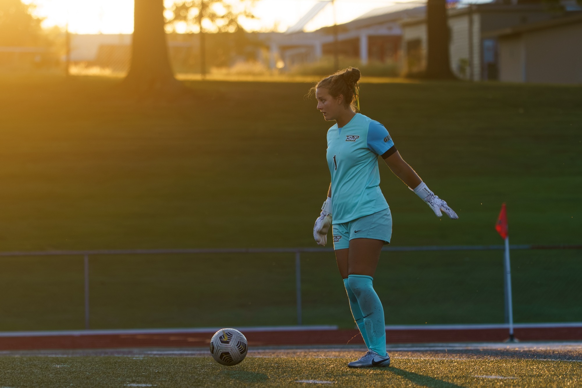 Catie Riddle in a teal/blue jersey standing by the soccer ball mentally preparing to punt it. There is a gorgeous sunset in the trees behind her.