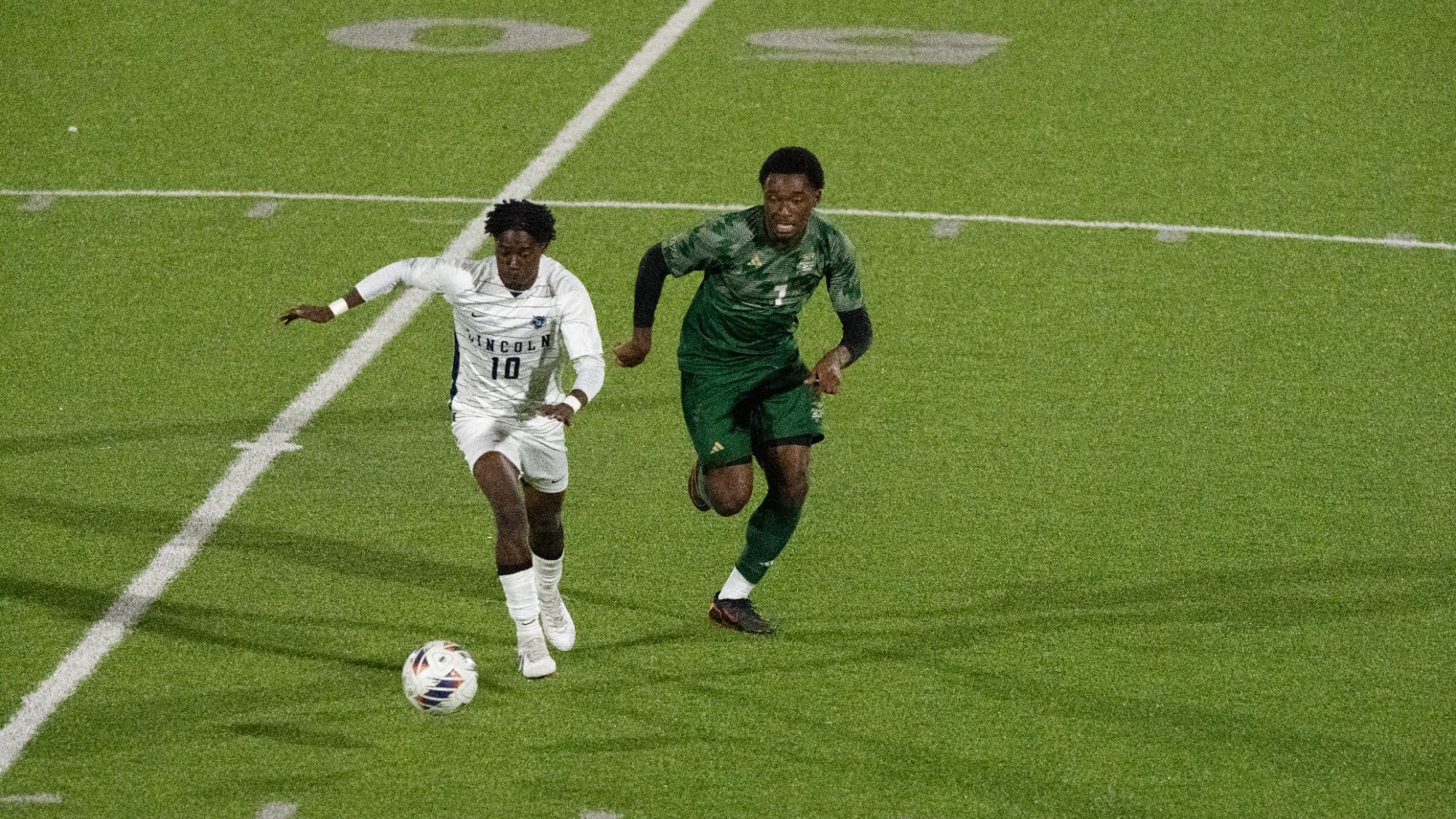 KhiBa Romany in a green jersey with a white 7 on the front and green shorts running after a Lincoln player in a white jeresey with a black number 10 across the front and white shorts who is following after the ball in front of him.