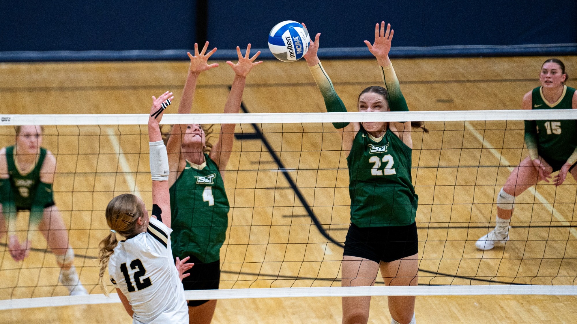Addie Schoenberger and Madison Smith of Missouri S&T Women's Volleyball photographed wearing green jerseys and jumping to block an attack during a match at Hammond Sports Complex