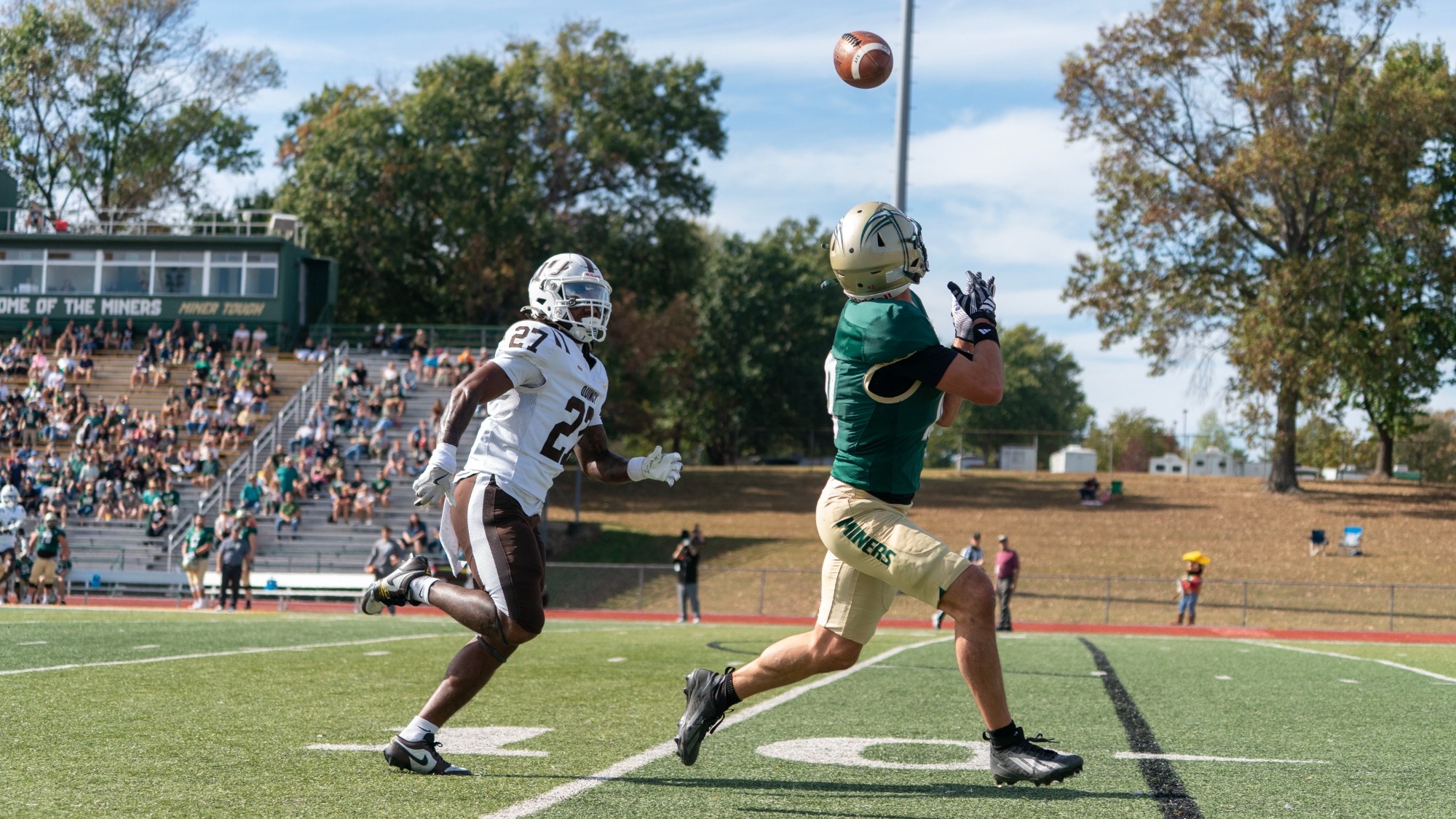 Tyler Gioia photographed wearing a green and gold football uniform with a gold helmet and being chased by a Quincy defender wearing white and brown. He is extending his arms to catch a pass during a game at Allgood-Bailey Stadium