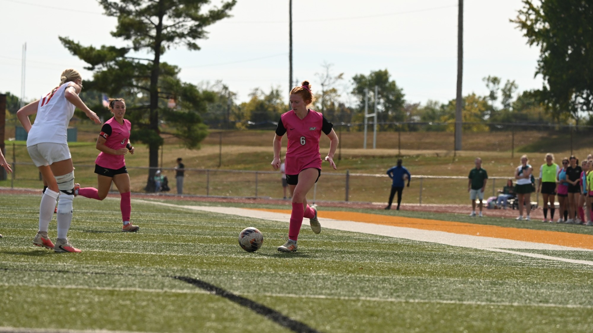 Aubrey Case in a pink jersey and pink shorts with a white number 6 on the chest dribbling the soccer ball away from a UMSL defender in a white jersey and white shorts. 
