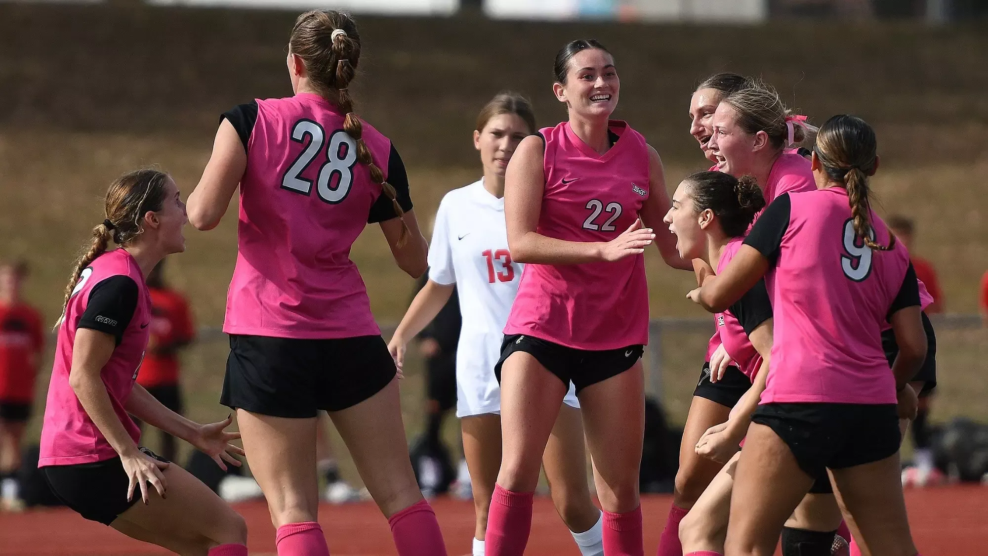 A group of Missouri S&T women's soccer players celebrate together on the field. The players wear pink jerseys with black sleeves, black shorts and pink socks. The team celebrates on a green artificial turf field with a red track and a grassy hillside in the background.