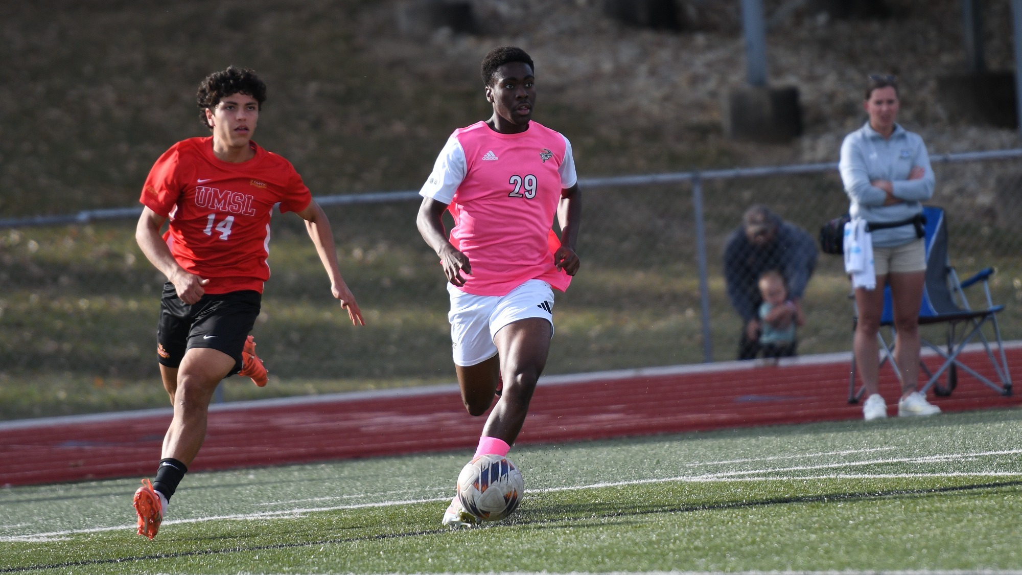 Eddy Kouadio in a pink S&T jersey with a white number 29 across the chest and pink shorts. He's dribbling the soccer ball away from an UMSL player in a red jersey and red shorts.