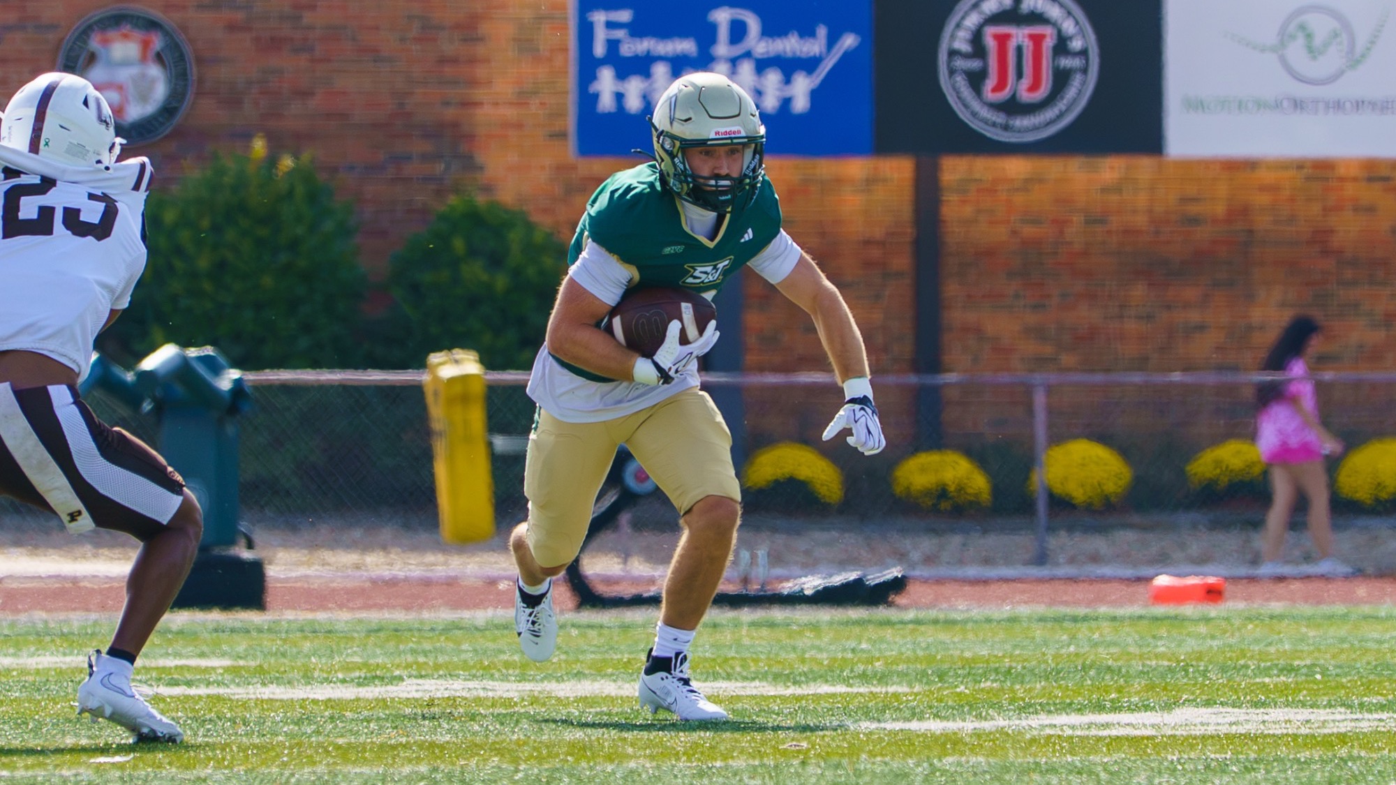 Reid Weber of Missouri S&T football photographed wearing a green and gold uniform and gold helmet, running while holding the football during a game in Allgood-Bailey Stadium