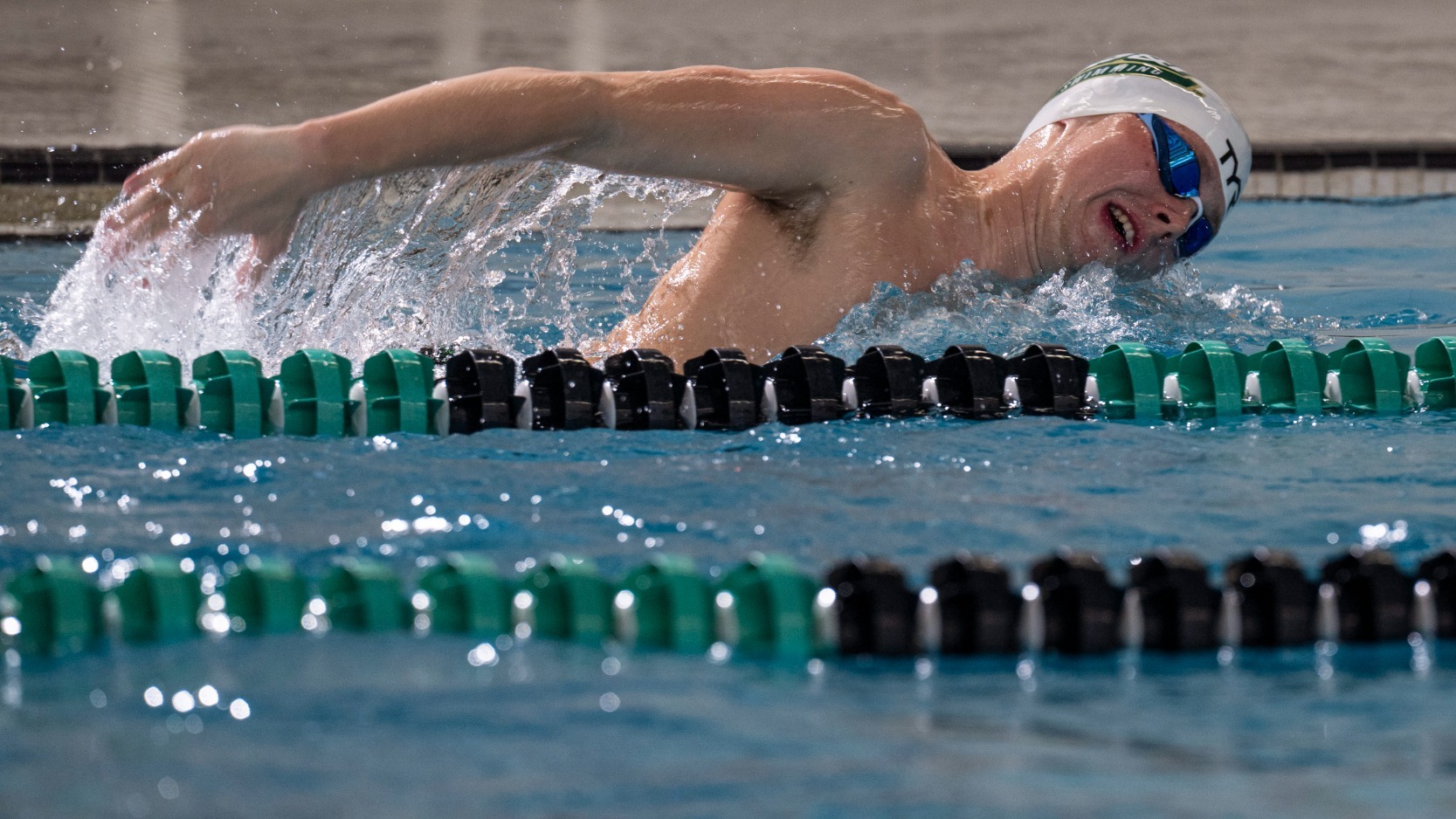 John Cottrell of Missouri S&T Swimming photographed during a freestyle event swimming in a pool while wearing a white cap that has the S&T Miners logo on the side