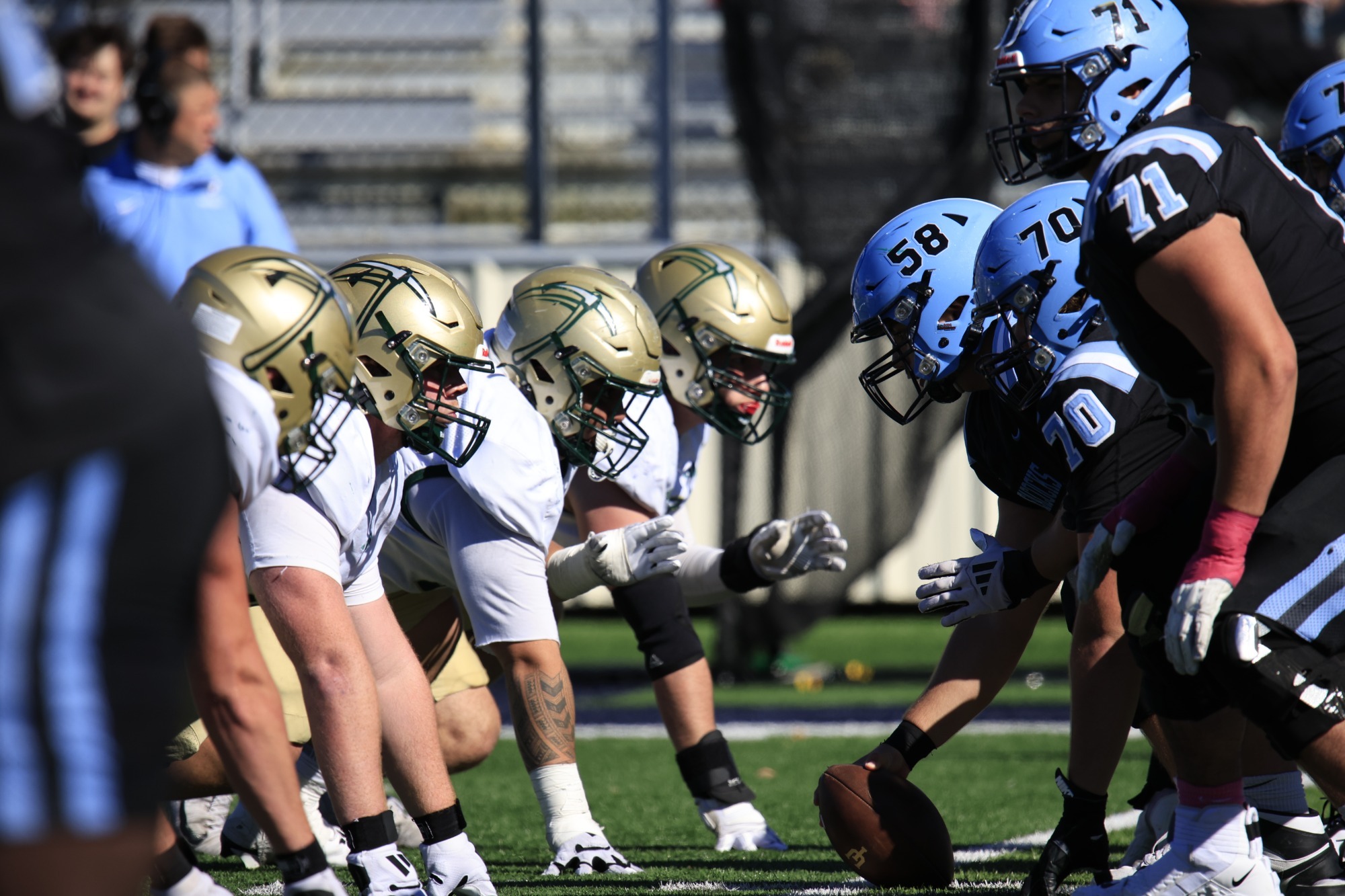 **Alt text:** Missouri S&T and Upper Iowa football players line up at the line of scrimmage before a play. Miners in white jerseys and gold helmets face Upper Iowa players in black jerseys and blue helmets on the field.