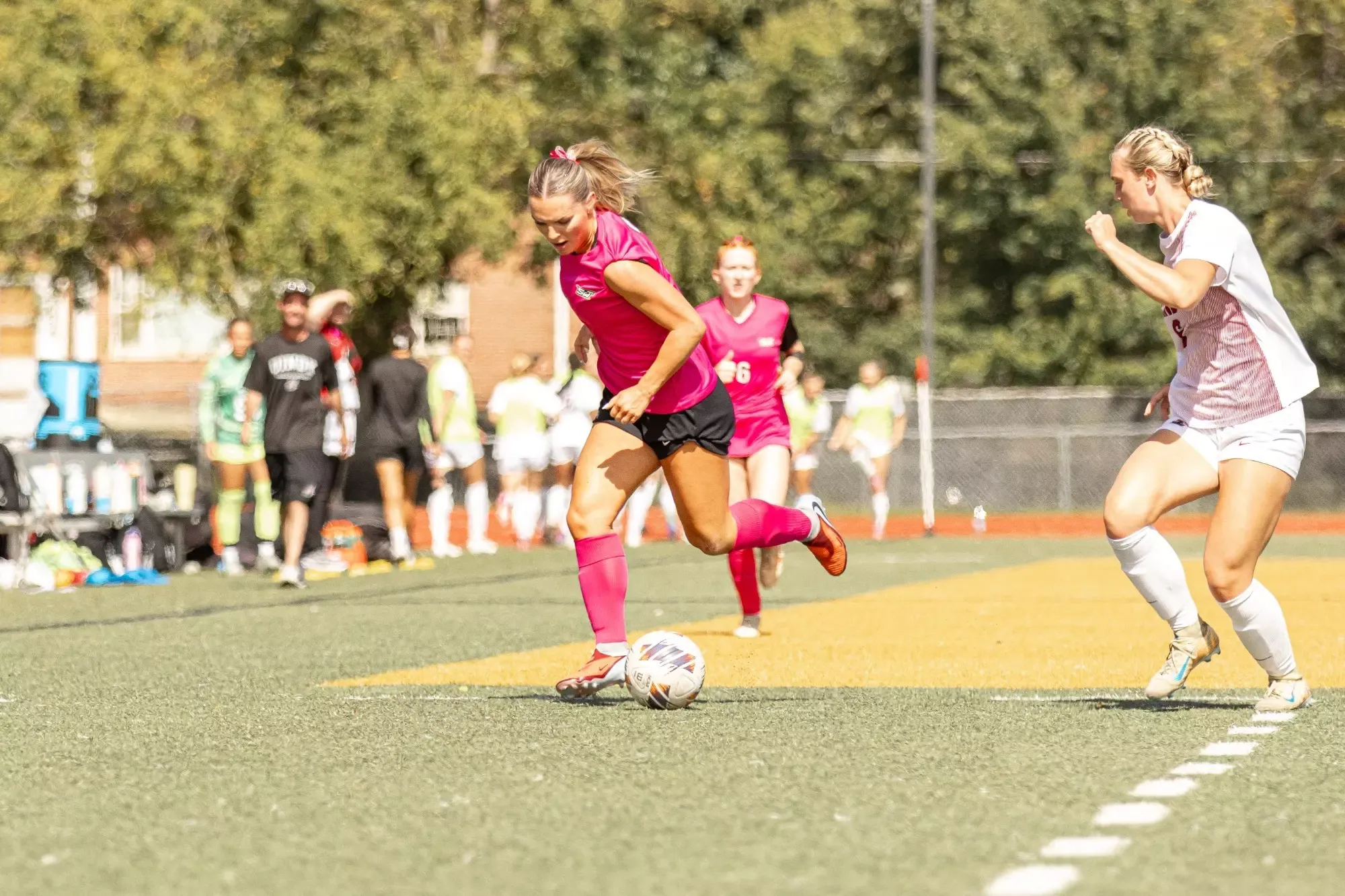 Missouri S&T women's soccer player No. 3 Jenna Marti dribbles the ball down the field during a match, pushing forward with a defender from the opposing team running alongside her. Marti wears a pink Missouri S&T jersey, black shorts and pink socks.