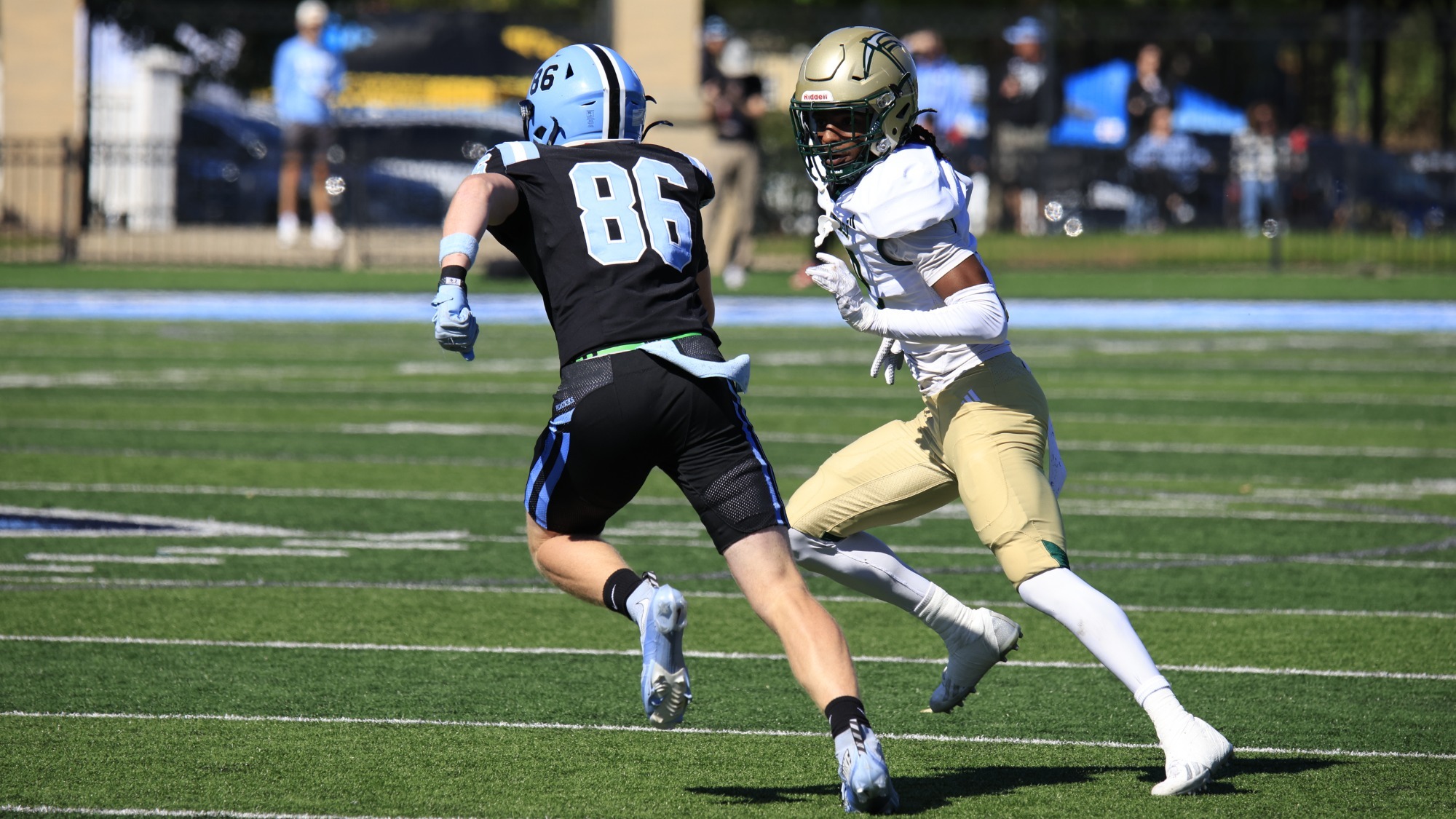 Kam Richardson, a Missouri S&T defensive back, runs across the field in a white jersey and gold pants while facing an opposing player wearing No. 86 in black.