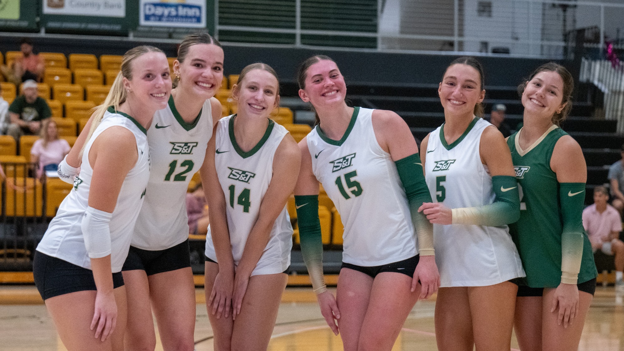 Maggie Miller, Dani Wilke, Peyton Svendsen, Elly Decker, Chelsea Plunkett, and Adelyn Weiler photographed posing together in white and green volleyball uniforms during a match in Gibson Arena