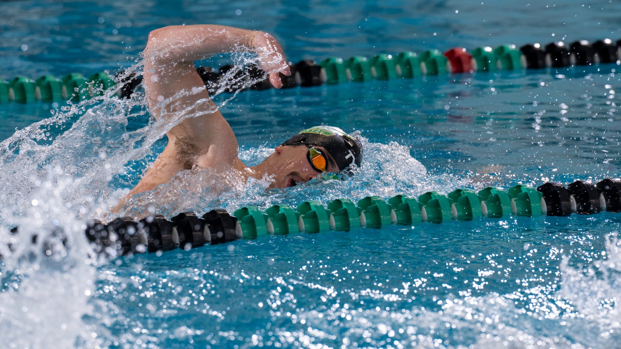 Eli Wetzel, a Missouri S&T men’s swimming athlete, is photographed swimming freestyle during a race in an indoor pool. He wears a Missouri S&T swim cap and goggles as his right arm recovers above the water and his face turns to the side to breathe. Water splashes around him as he moves down the lane, with green and black lane lines visible on either side.