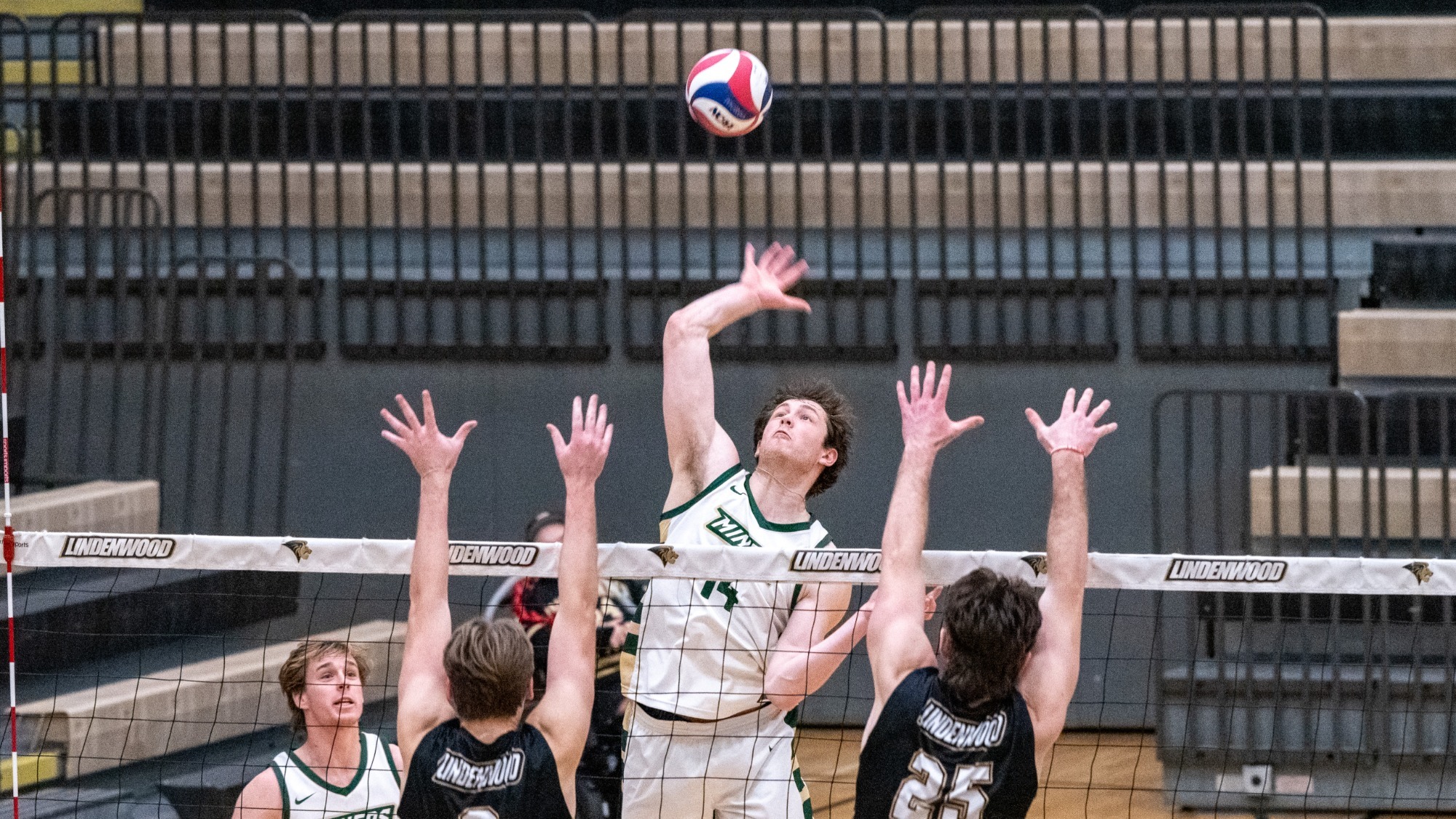Aaron Sallade, a Missouri S&T men’s volleyball player, is photographed jumping at the net and reaching with his right arm to attack the ball during a match. He wears a white Missouri S&T uniform with green accents and is shown mid-air as the volleyball floats above the net. Two opposing players in black Lindenwood jerseys jump in front of him with both arms raised, attempting to block the attack. Teammates in white are positioned on either side of the play, watching the action.