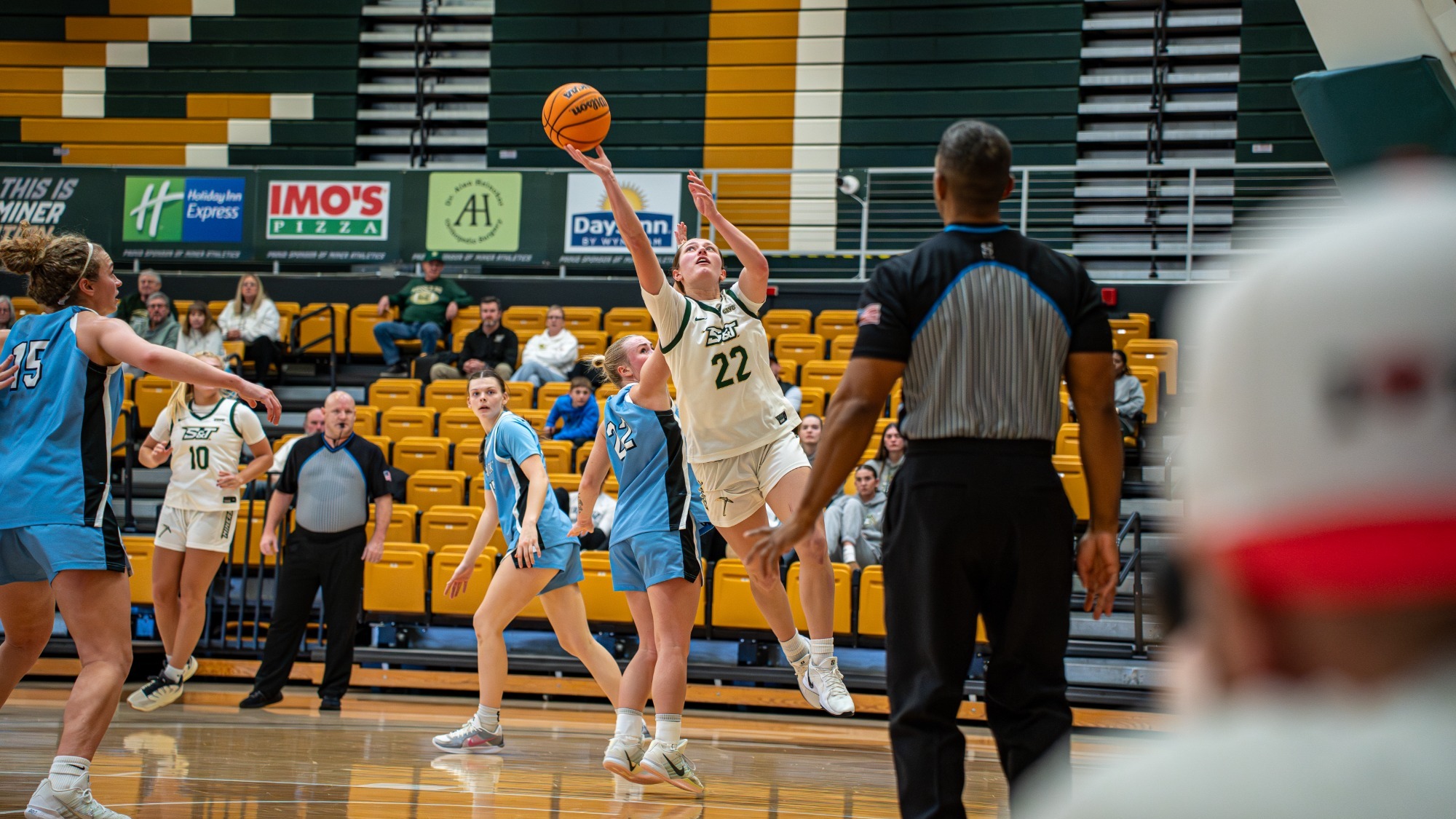 Missouri S&T guard Adriana Benassi in white mid layup with the ref infront ofe her and Upper Iowa defenders around her in blue.