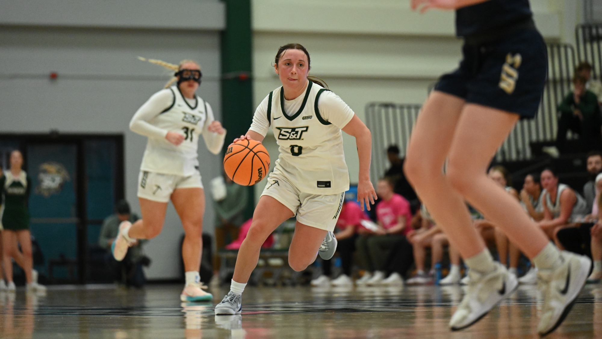 Missouri S&T women's basketball athlete Macy Marraccini wearing a white jersey while dribbling the basketball with her right hand with a UIS defender blurred in the foreground and a teammate and fans visible in the background during a game in Gibson Arena