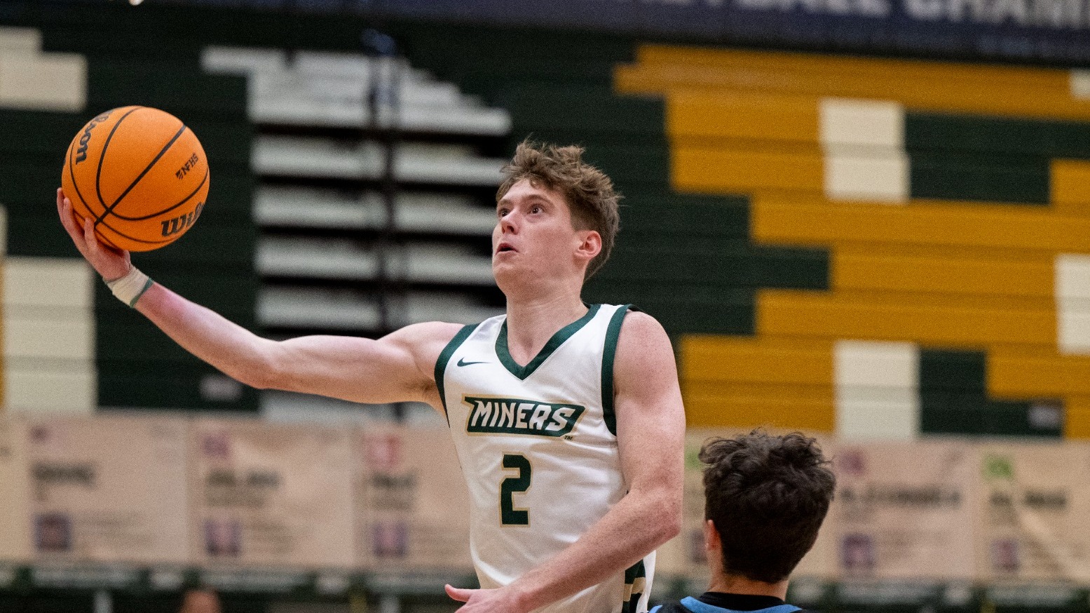 Missouri S&T men's basketball athlete Alex Benassi wearing a white jersey and holding out the basketball with his right arm as he goes for a layup during a game in Gibson Arena