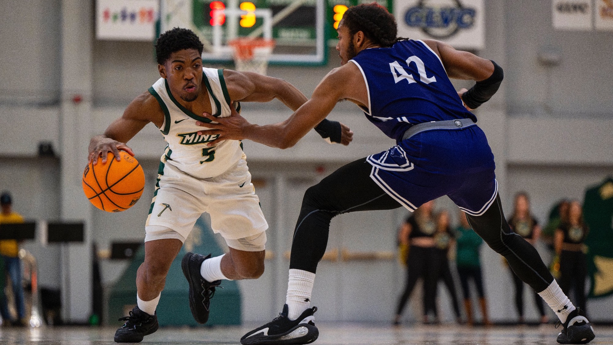 Missouri S&T men’s basketball athlete Darron Boodah Henry wearing a white jersey while dribbling the basketball with his right hand in an attempt to dribble around a McKendree defender. The McKendree defender is wearing a purple uniform and has his hand out to prevent Henry from getting by.