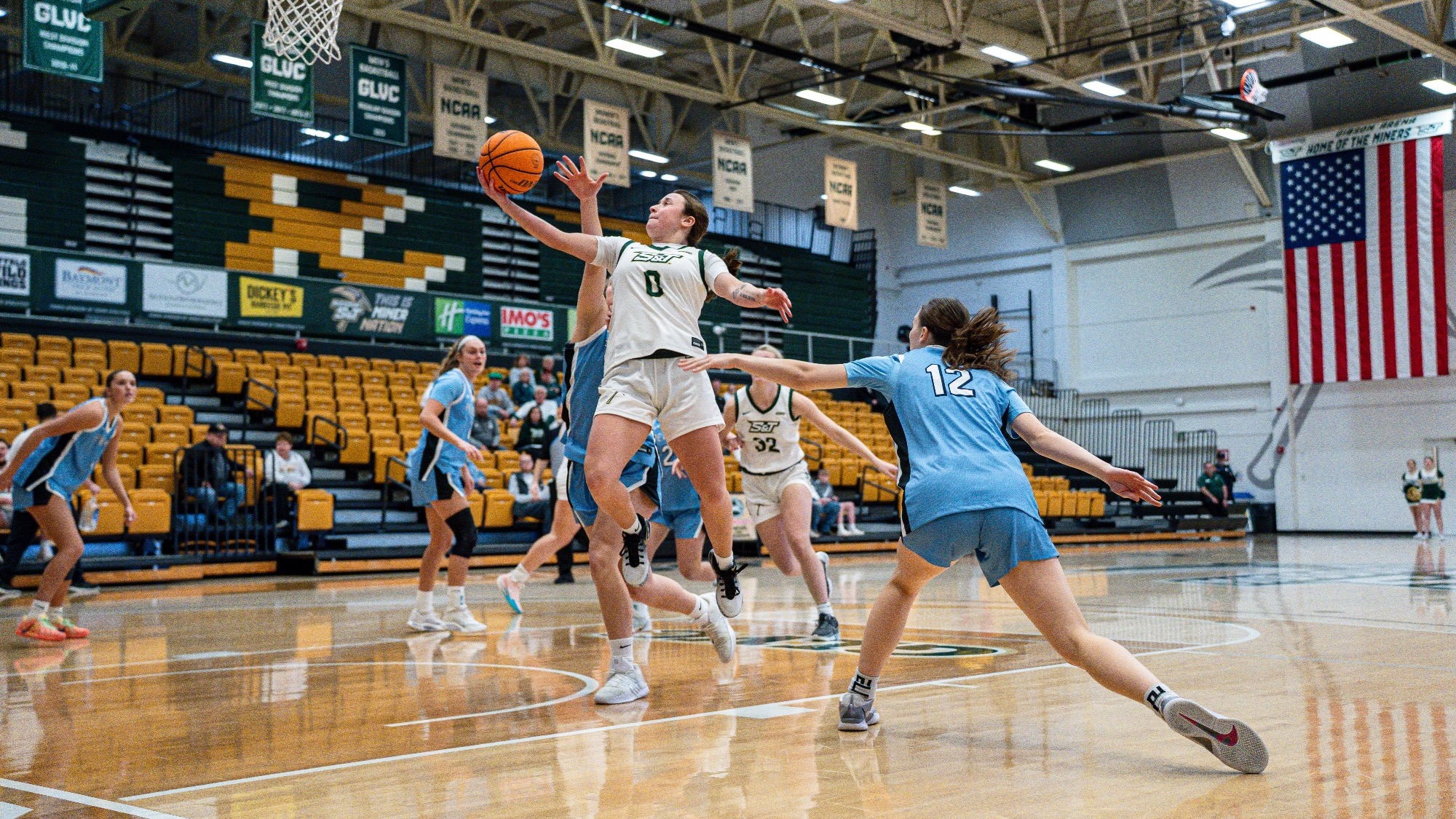 Missouri S&T women's basketball player Macy Marraccini in a white S&T jersey going up for a layup with Upper Iowa players in baby blue jersey around her.