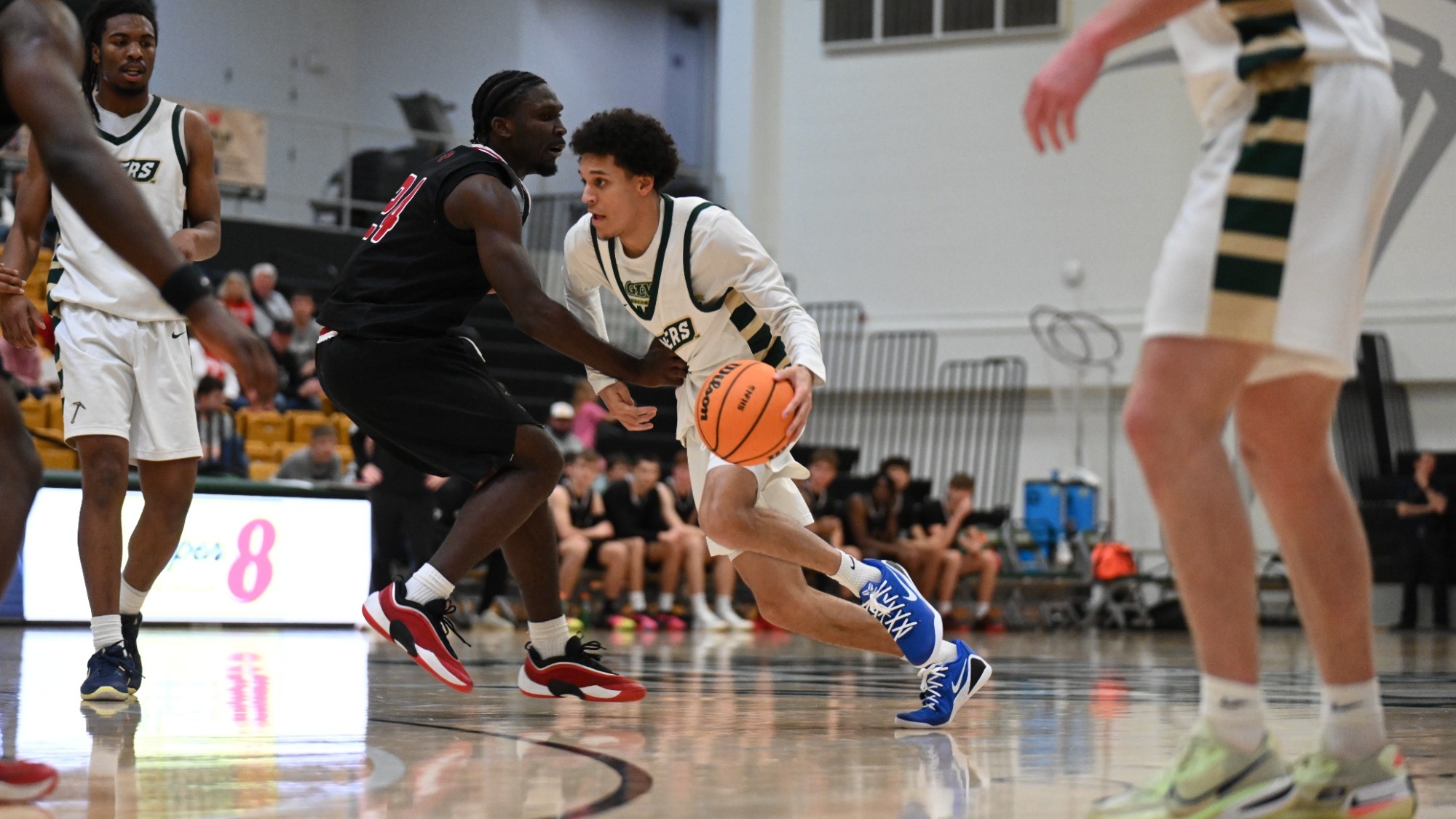 Blaise Beauchamp is photographed driving to the basket during a basketball game, dribbling with his right hand as he leans past a defender in a black uniform. He wears a white jersey with green trim and bright blue shoes, with teammates and the bench visible in the background inside the gymnasium.