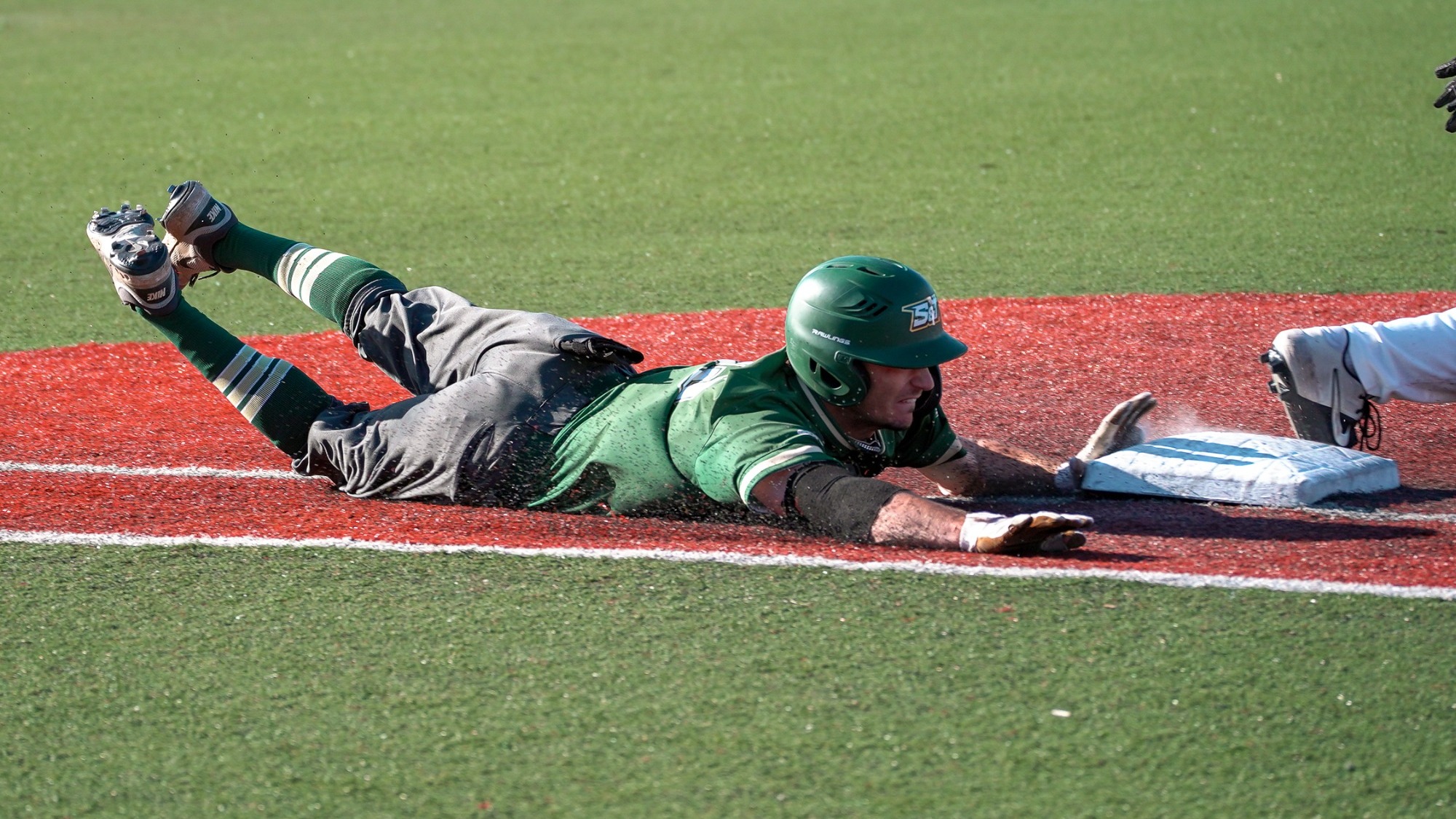 Garrett Meyer is photographed diving headfirst toward a base on a red turf infield, fully extended with his arms outstretched and cleats lifted off the ground. He wears a green jersey and helmet with gray pants, as dirt pellets spray up around him while he reaches for the bag during a close play.