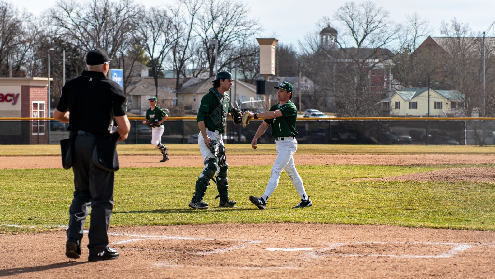 Tyler Bizzle, a Missouri S&T catcher, is photographed in full green catching gear meeting pitcher Andrew Hedgecorth, who is wearing a green jersey and white pants, on the infield grass during a game. An umpire stands near home plate in the foreground, while additional Miners players are visible in the background near the mound and outfield fence on a sunny afternoon.