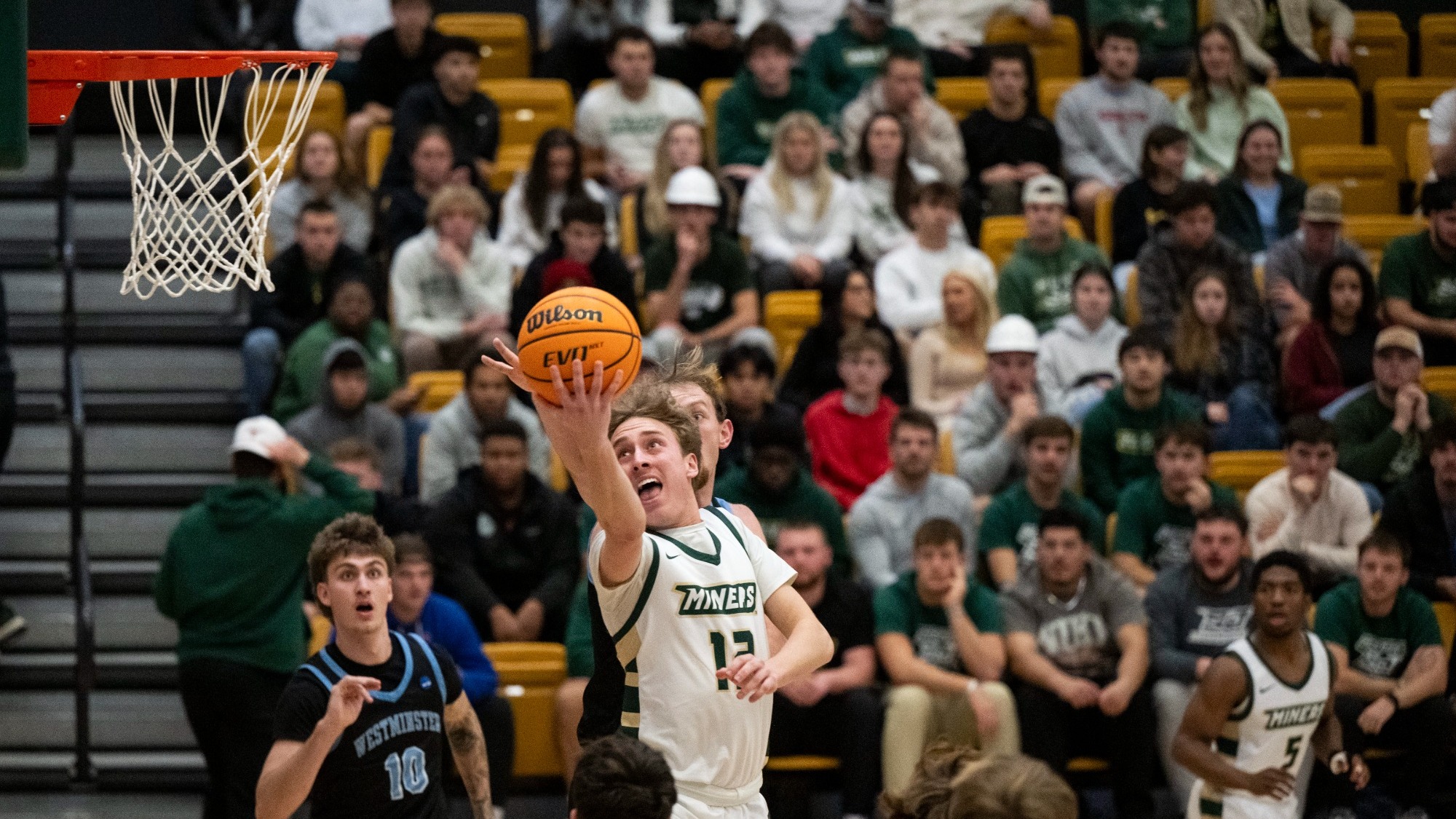 Missouri S&T men's basketball athlete Aidan Burns wearing a white S&T jersey and reaching out with his right arm for a reverse layup with the goal visible to the right, with fans and opposing players visible in the background.